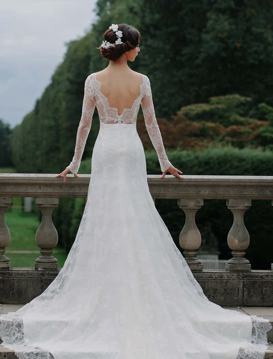 Wedding Gown 桌布 壁紙 — 一位身穿白色蕾絲長袖婚紗的新娘站在石造陽台上，俯瞰著鬱鬱蔥蔥的綠植。