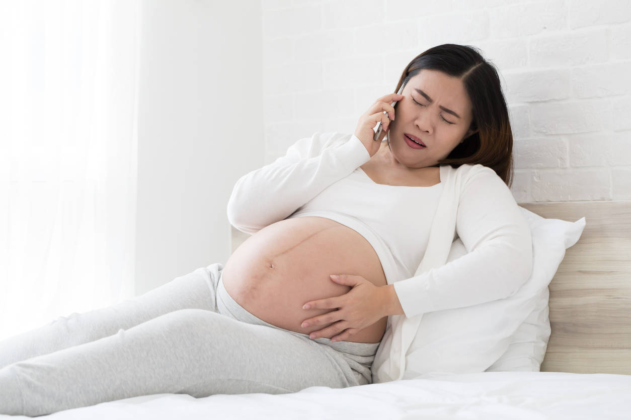 Pregnant 桌布 壁紙 — 一位身穿白色衣服的孕婦躺在床上，抱著肚子，表情痛苦地講電話。