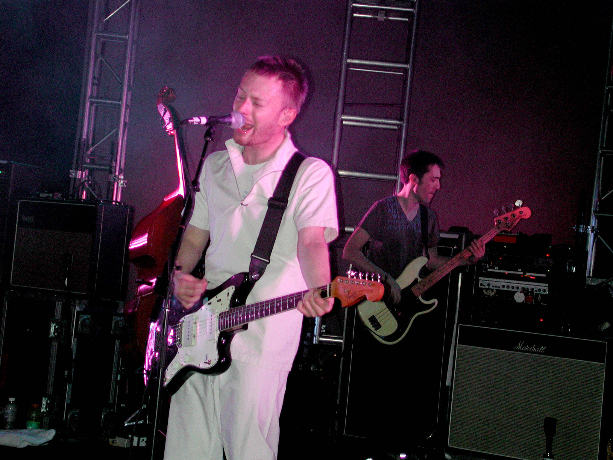 band 桌布 壁紙 — 一位身穿白色衣服的音樂家在粉紅色舞台燈光下彈奏電吉他，另一位吉他手在他身後彈奏。