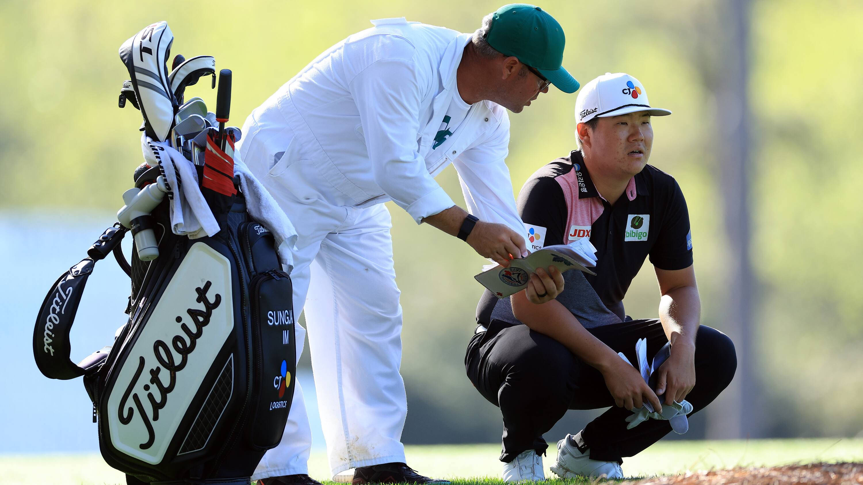 運動 桌布 壁紙 — 一位身穿白色衣服的高爾夫教練彎下腰，與蹲在 Titleist 球袋旁的高爾夫球手在陽光明媚的球場上討論策略。