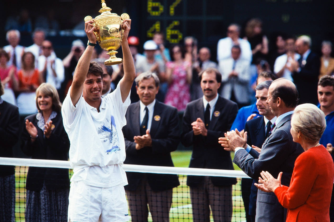 Tennis 桌布 壁紙 — 一位身穿白色裝備的勝利網球選手在草地場上將金色獎盃高舉過頭，觀眾和官員在周圍鼓掌。
