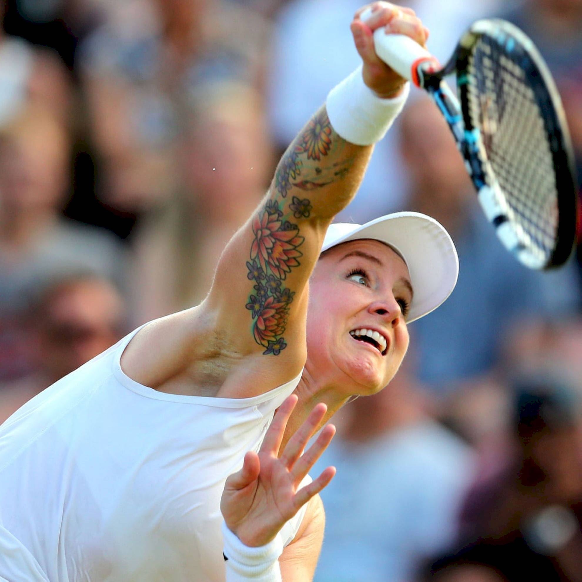 Tennis 桌布 壁紙 — 一位身穿白色裝備的女網球運動員，高舉帶有花卉刺青的手臂揮拍，露出燦爛的笑容。