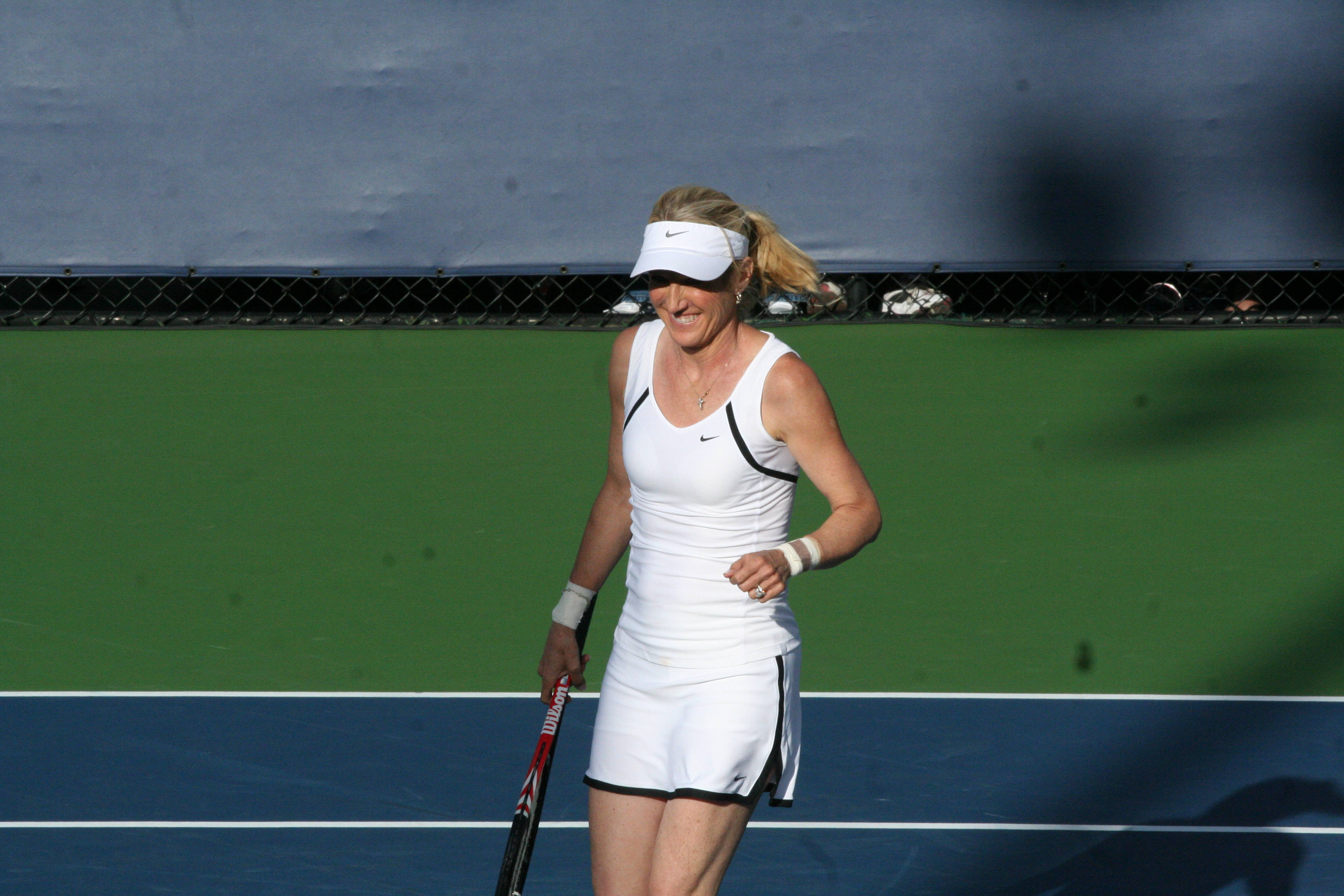 Tennis 桌布 壁紙 — 一位身穿白色裝備的女網球運動員在藍綠色球場上微笑著拿著球拍。