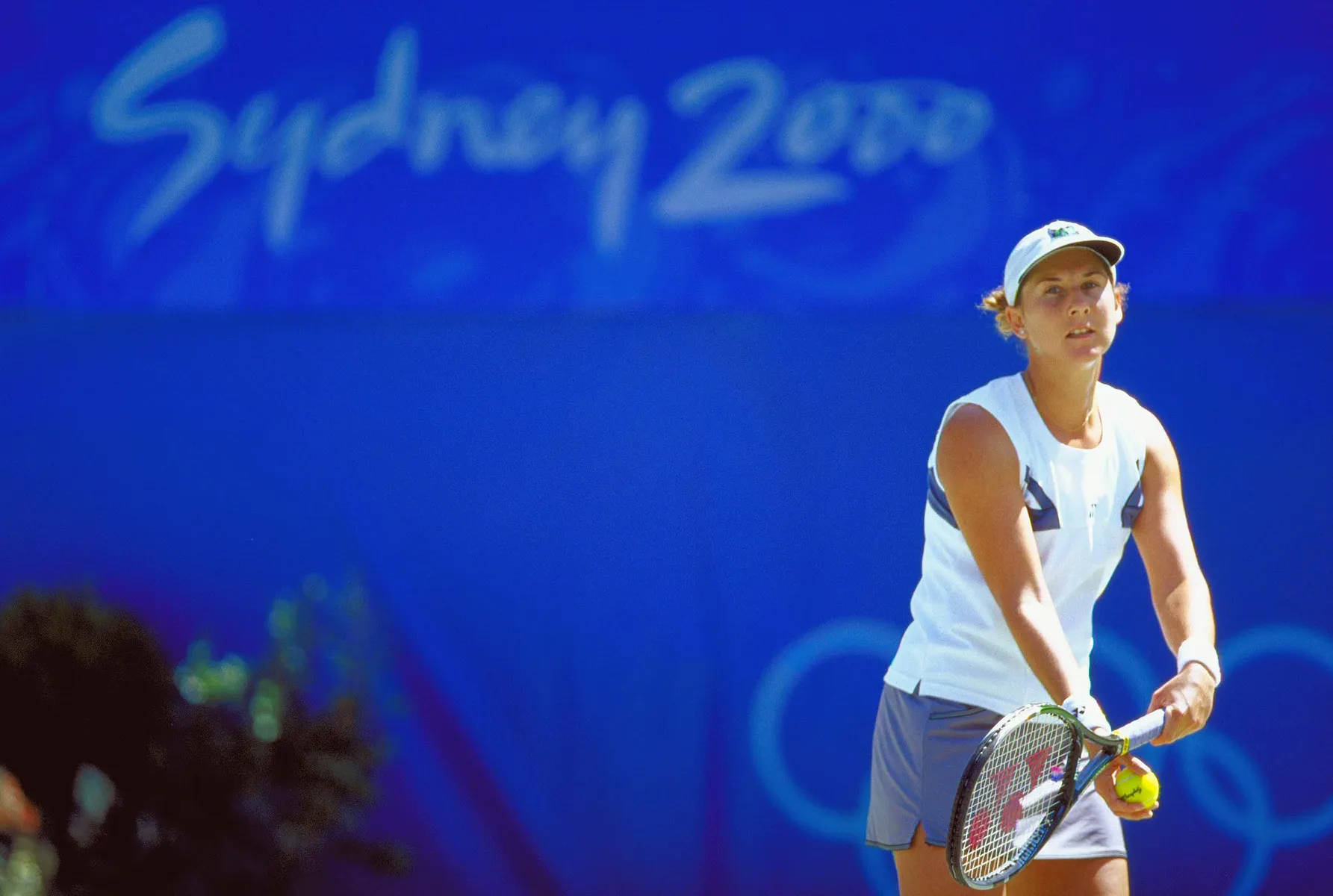 Monica Seles 桌布 壁紙 — 一位身穿白色裝備的女網球選手在充滿活力的藍色背景下準備在雪梨2000奧運會上發球。