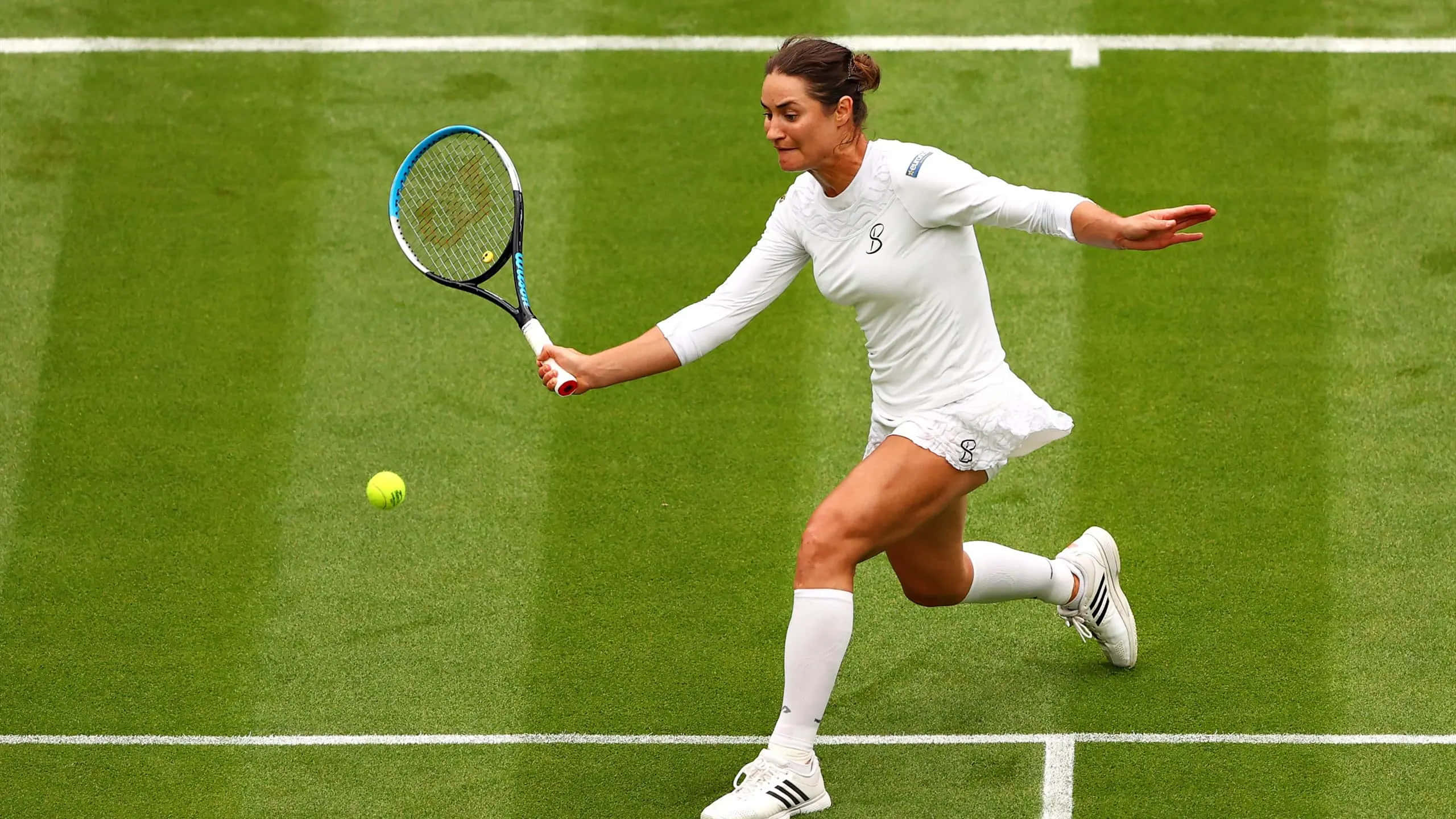 Tennis 桌布 壁紙 — 一位身穿白色裝備的女網球選手在綠色草地場上向前衝刺，用球拍擊打黃色球。