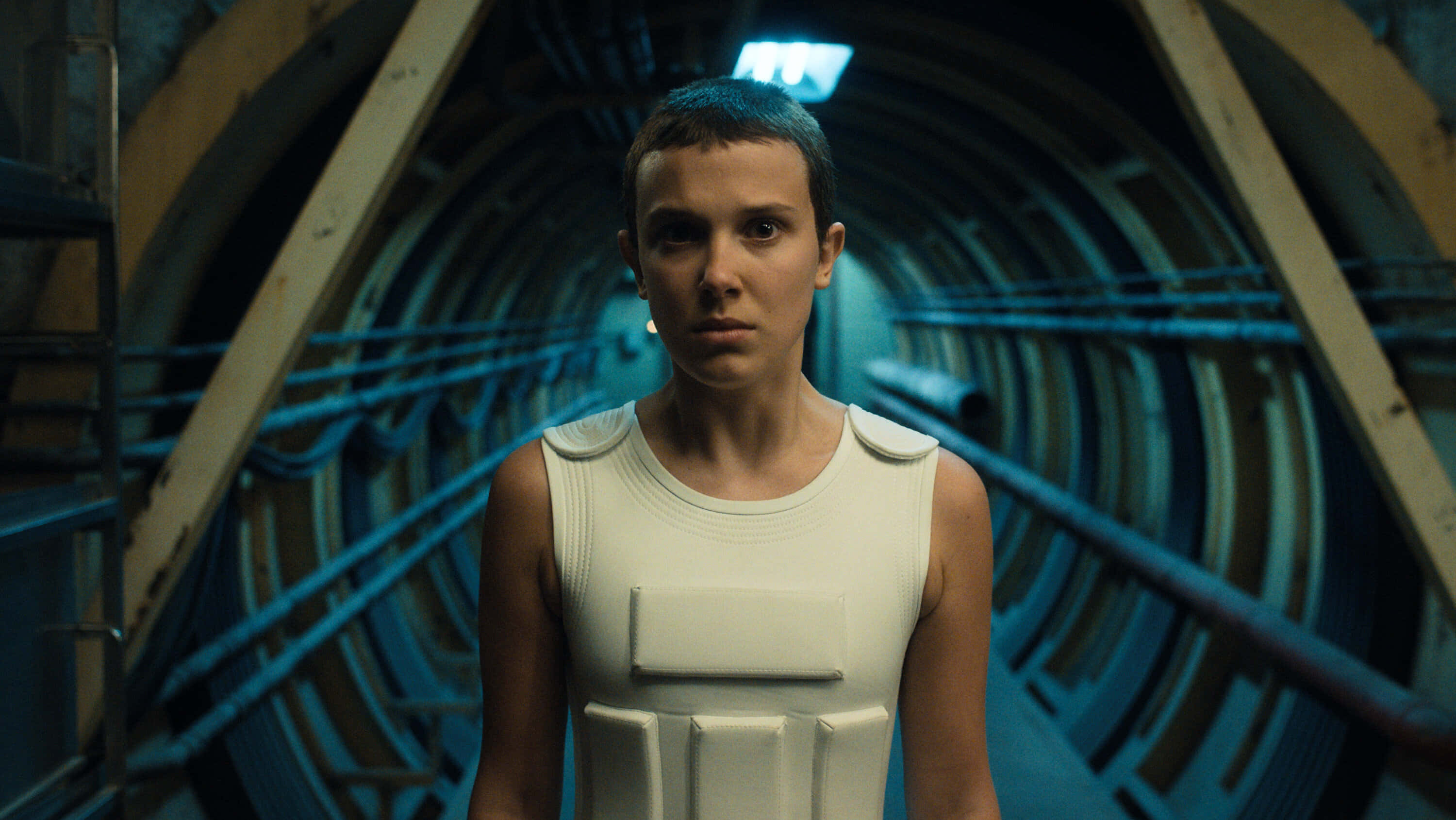 Eleven Stranger Things 桌布 壁紙 — 一位身穿白色裝甲背心的年輕女子，莊嚴地站在一個光線昏暗、帶有藍色照明的工業隧道中。