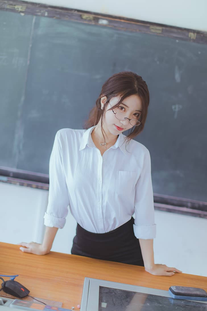  壁紙 — 一位身穿白色襯衫、戴眼鏡的年輕女子靠在教室的木桌上，身後是黑板。