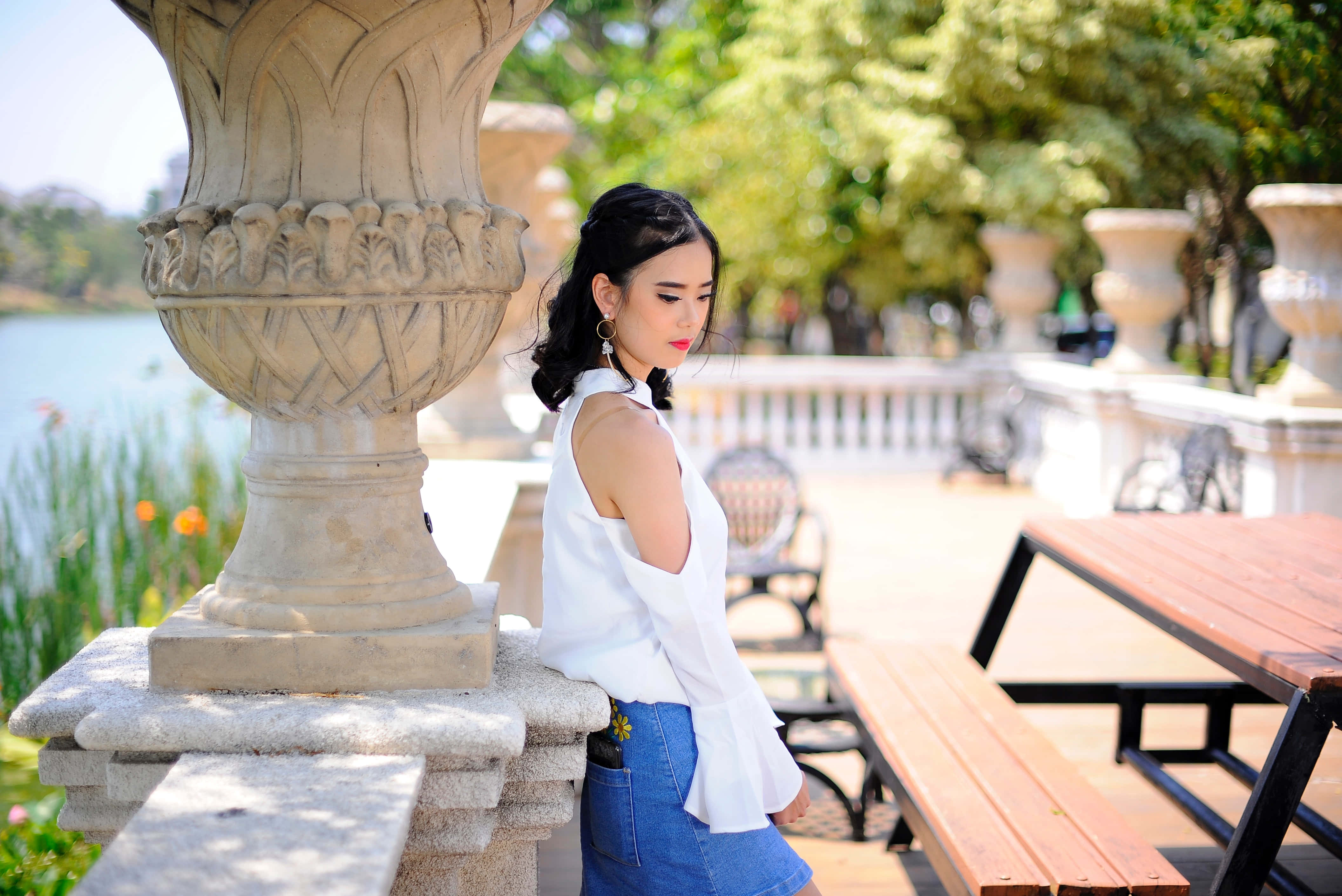 Summer Fashion 桌布 壁紙 — 一位身穿白色襯衫和牛仔裙的年輕女子在陽光明媚的公園裡靠著石柱，背景有湖泊和裝飾甕。