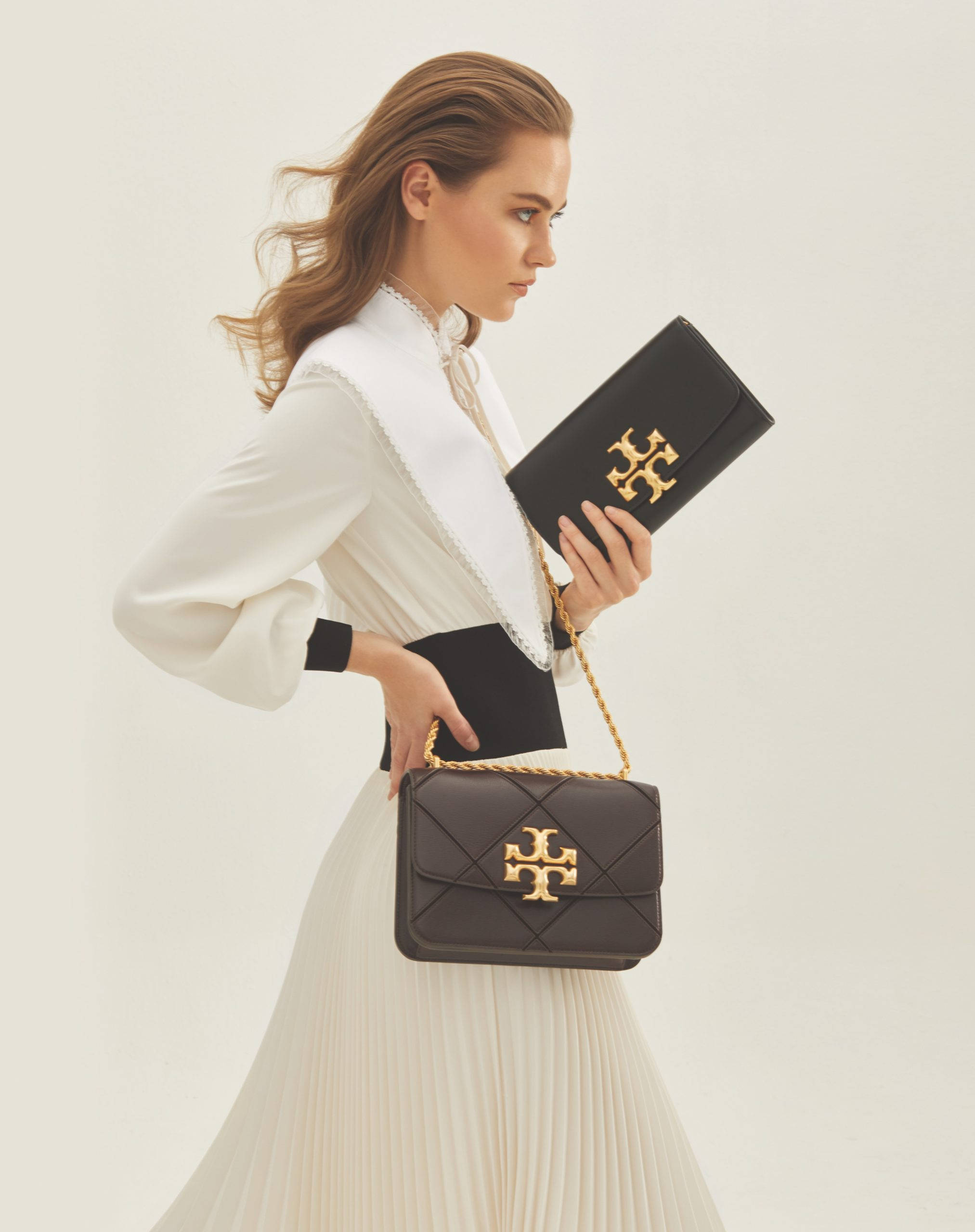 Tory Burch 桌布 壁紙 — 一位身穿白色襯衫和百褶裙的女性，在純色背景前拿著一個帶金色金屬配件的黑色絎縫手提包。
