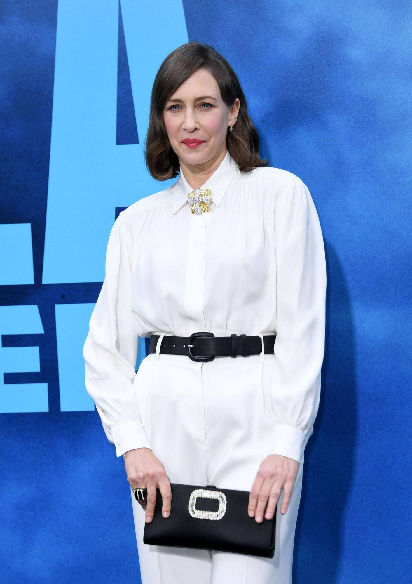 Celebrity 桌布 壁紙 — 一位身穿白色襯衫和長褲、搭配黑色腰帶和手拿包的女性，在鮮明的藍色背景前自信地站立。