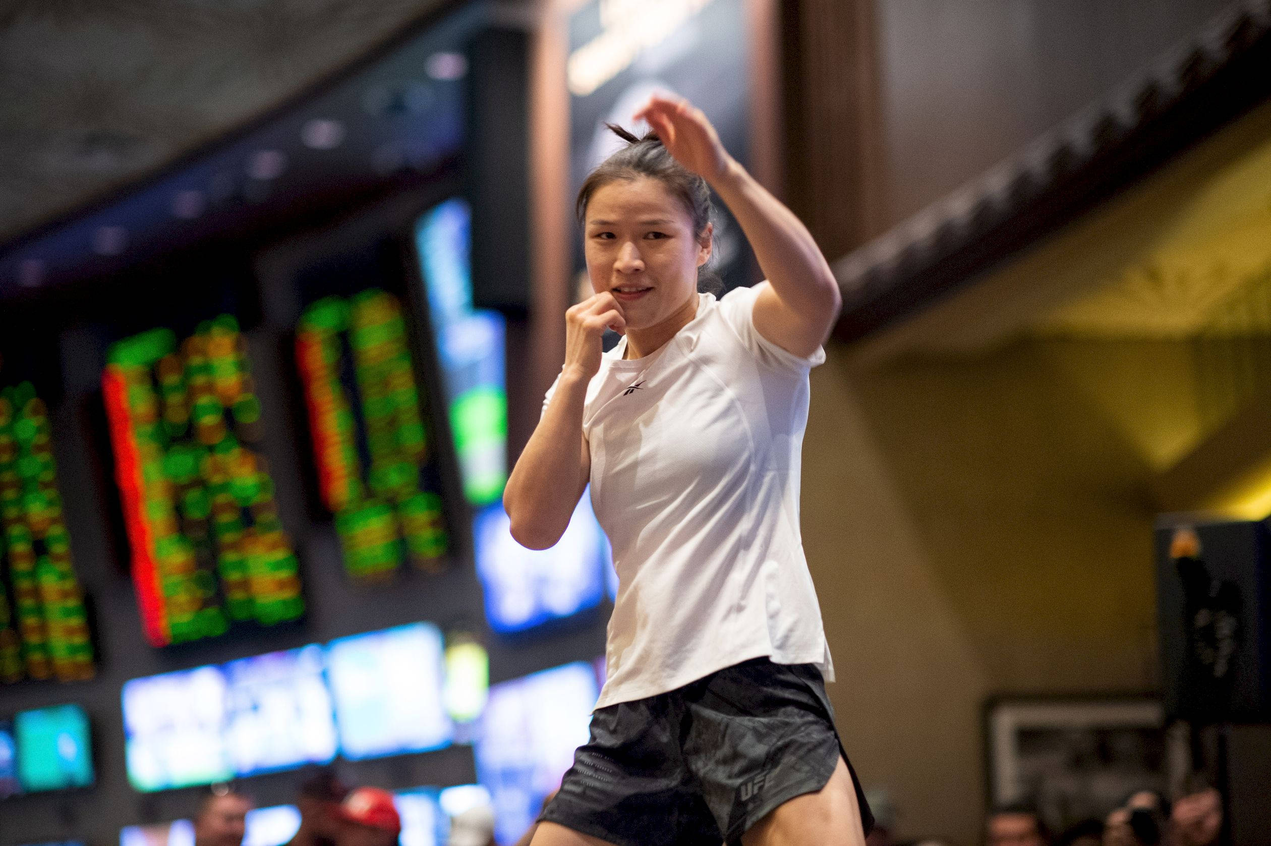 Athletic 桌布 壁紙 — 一位身穿白色襯衫和黑色短褲的女戰士，在昏暗的競技場中，身後有發光的數位計分板，擺出動態姿勢。