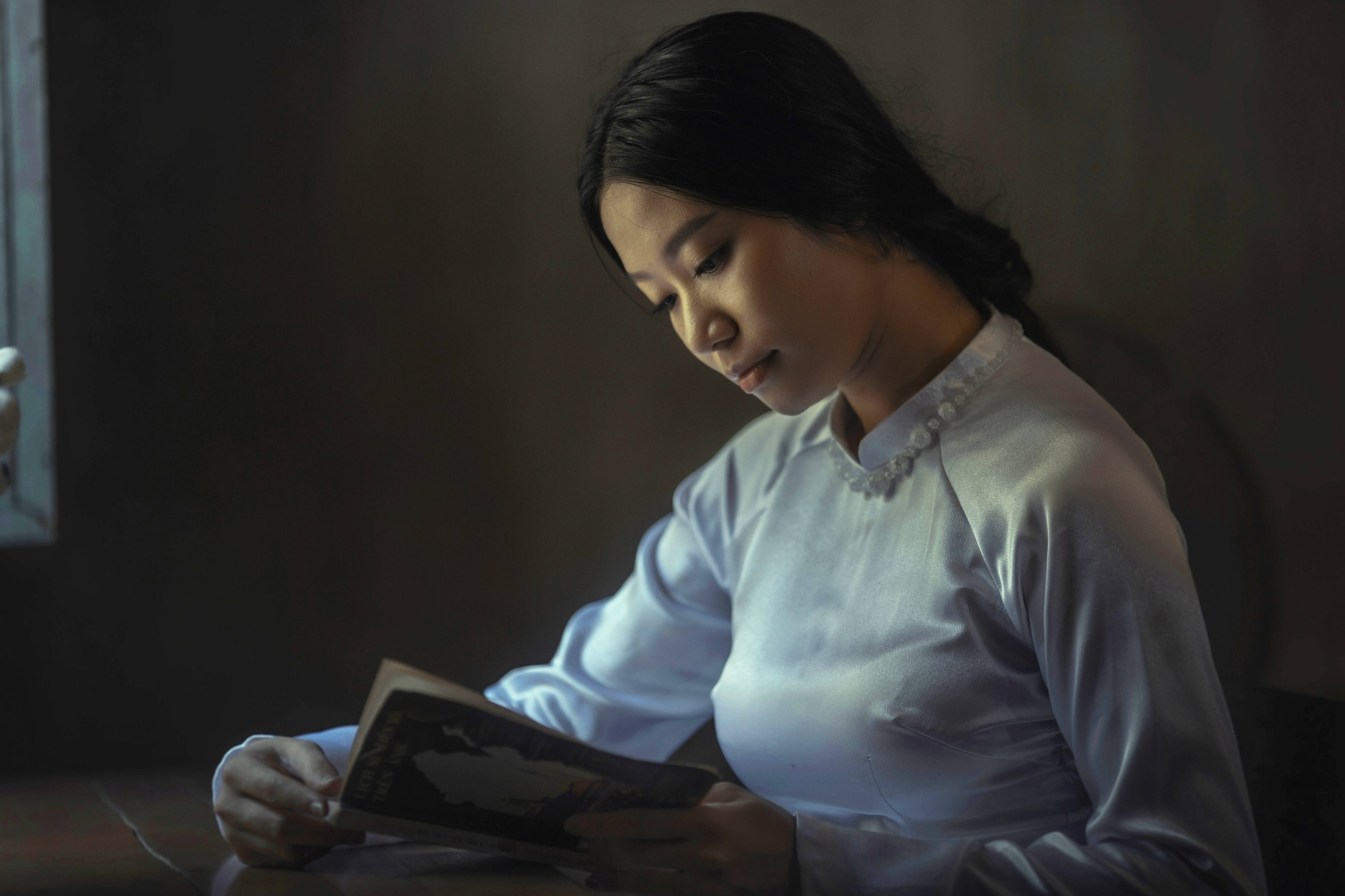 看書 桌布 壁紙 — 一位身穿白色襯衫的女性在昏暗的房間裡，藉著柔和窗光閱讀一本書。