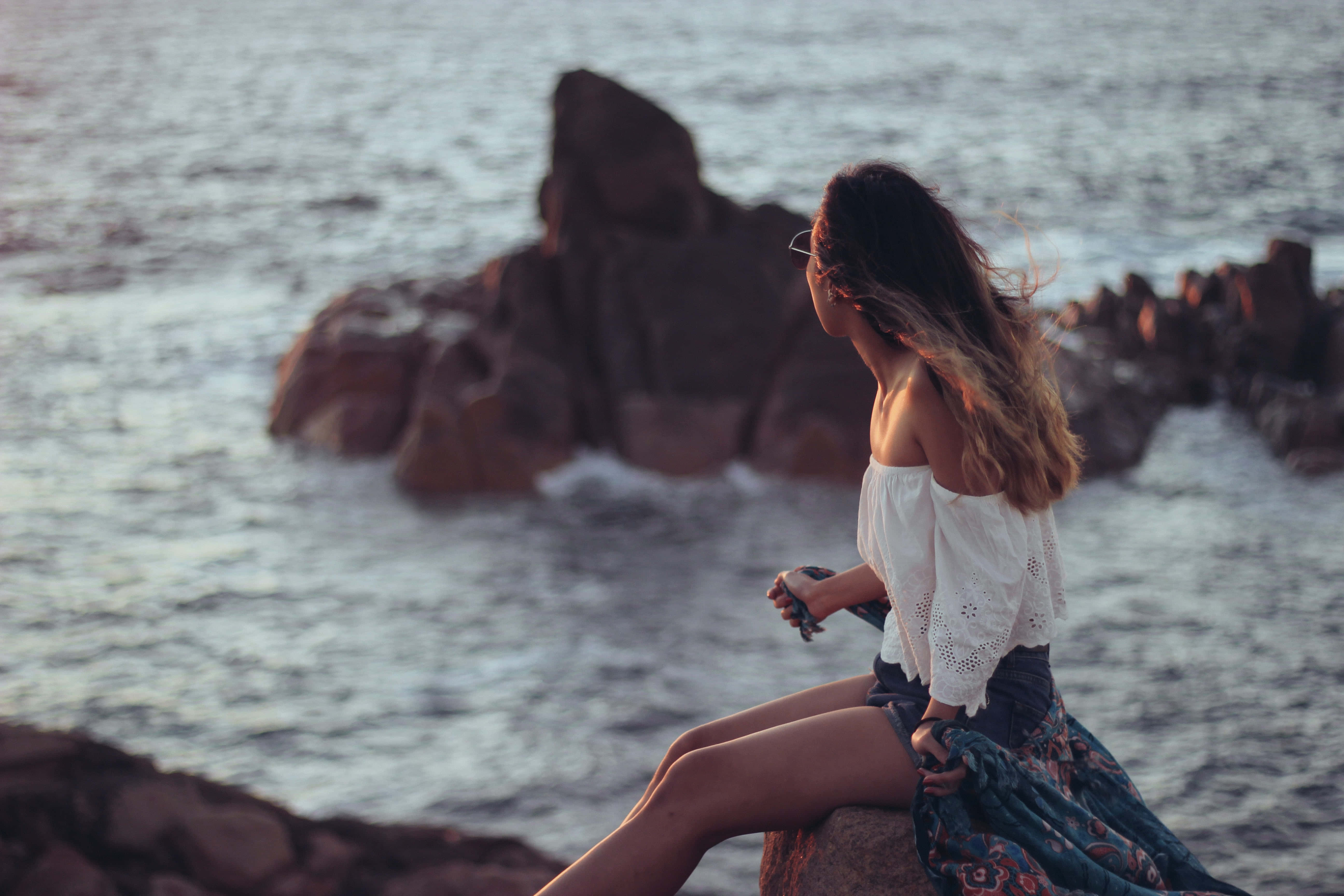 Summer Vacation 桌布 壁紙 — 一位身穿白色襯衫的女性坐在岩石懸崖上，俯瞰海洋，正值黃金時刻，風吹動她的頭髮。