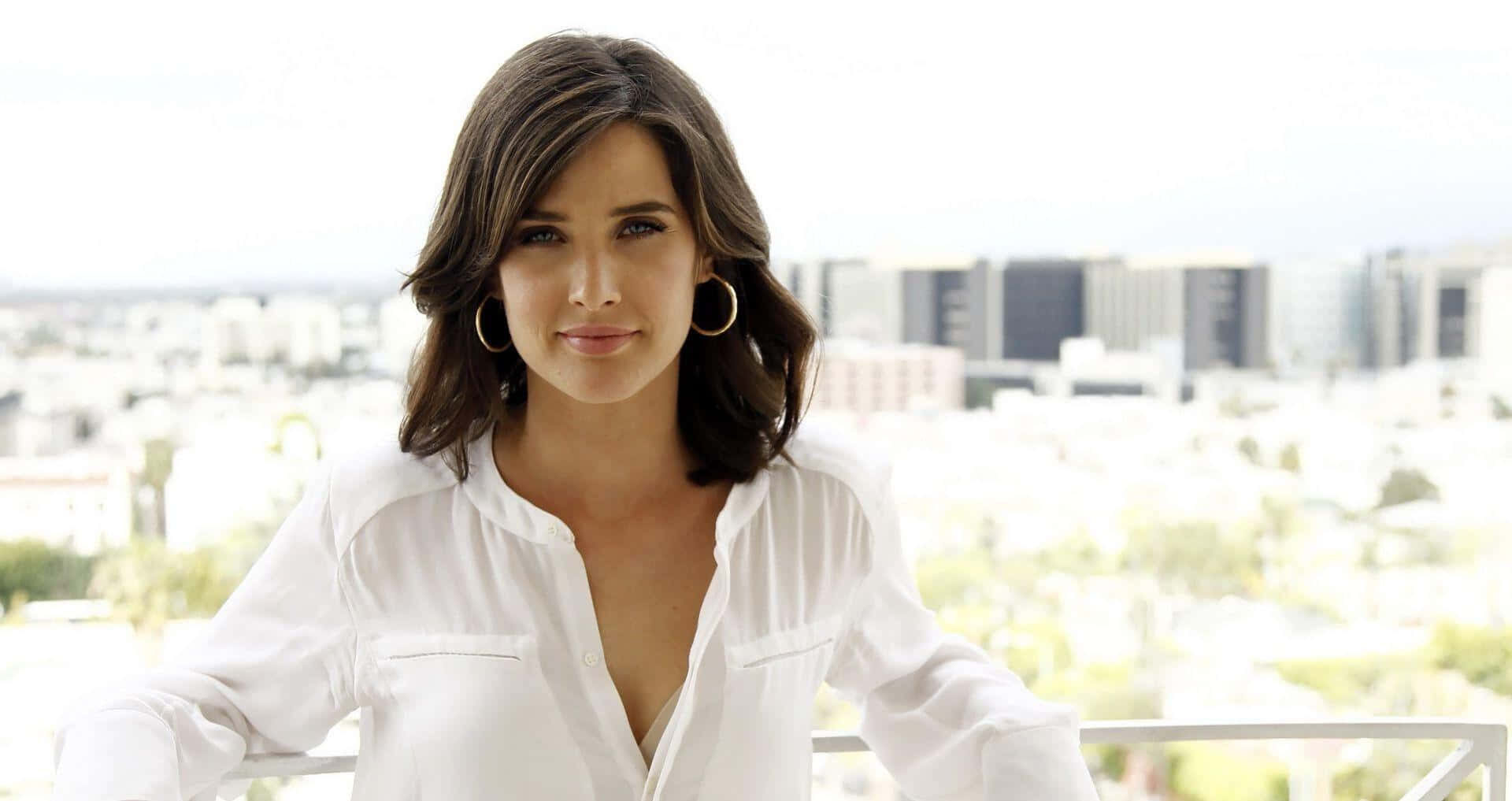 Cobie Smulders 桌布 壁紙 — 一位身穿白色襯衫的女性站在陽台上，俯瞰著充滿現代建築的明亮城市景觀。