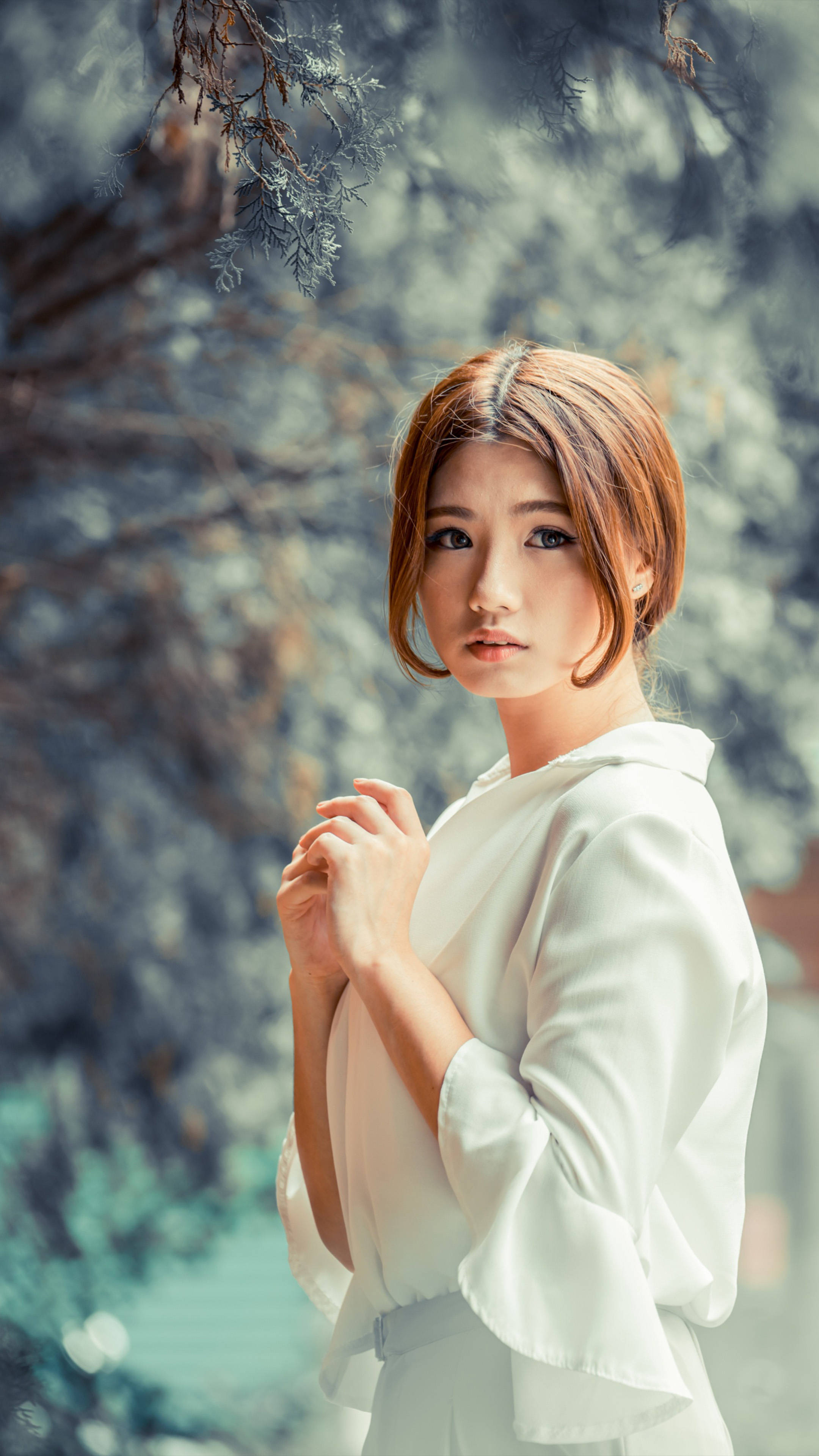 phone girl 桌布 壁紙 — 一位身穿白色襯衫的年輕女性在柔和的自然光下優雅地站在戶外，模糊的樹葉環繞著她。