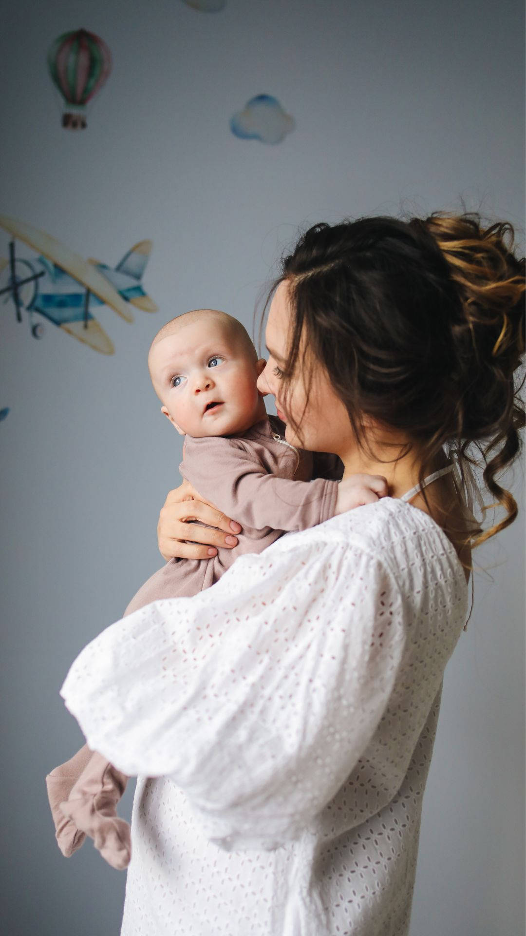Mom 桌布 壁紙 — 一位身穿白色襯衫的母親，溫柔地抱著她的寶寶，靠在一面裝飾有飛機和熱氣球貼紙的牆上。