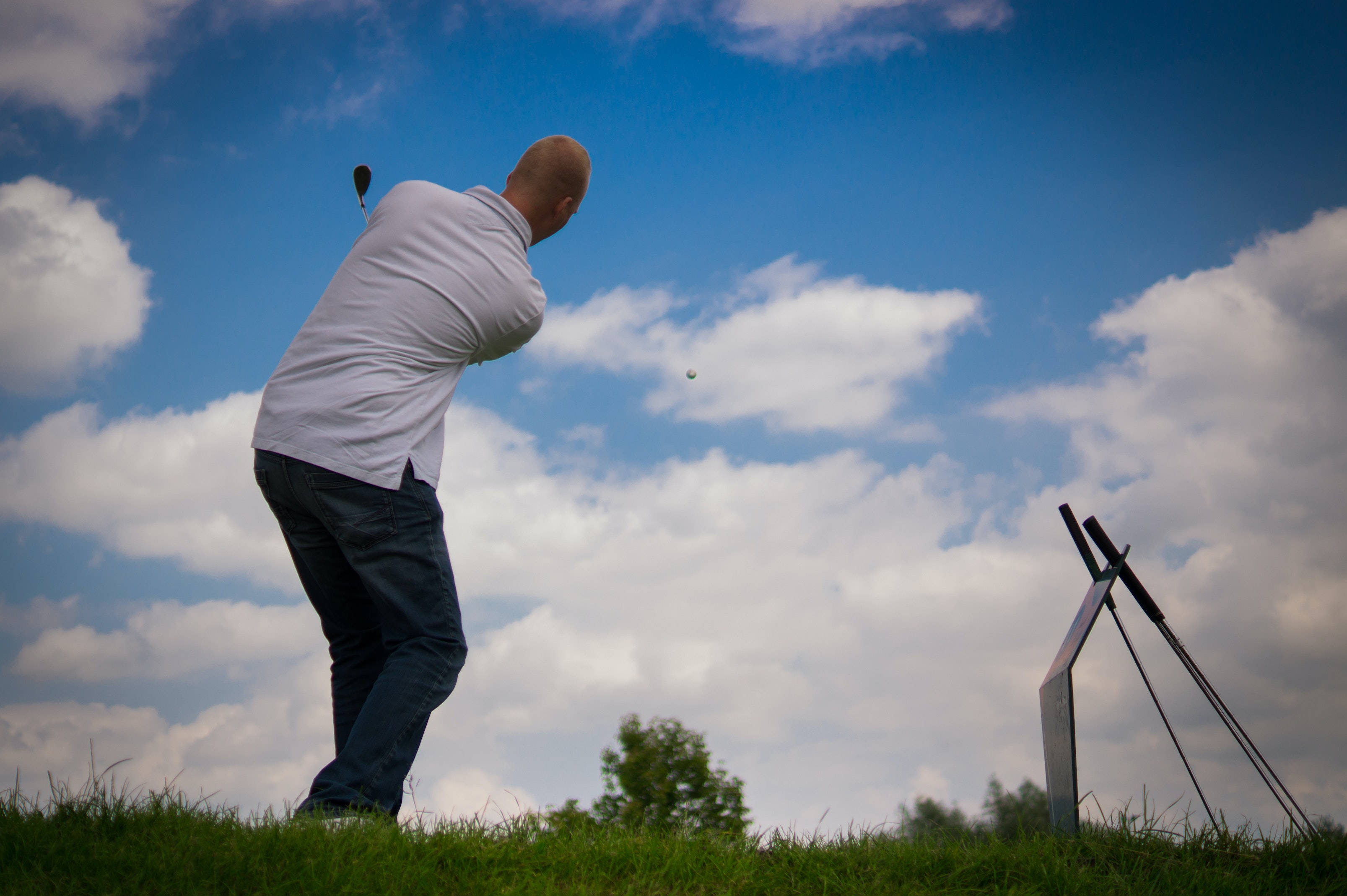 4K Golf 桌布 壁紙 — 一位身穿白色襯衫的高爾夫球手在明亮的藍天下揮桿，天空中有蓬鬆的白雲，下面是綠色的草地。