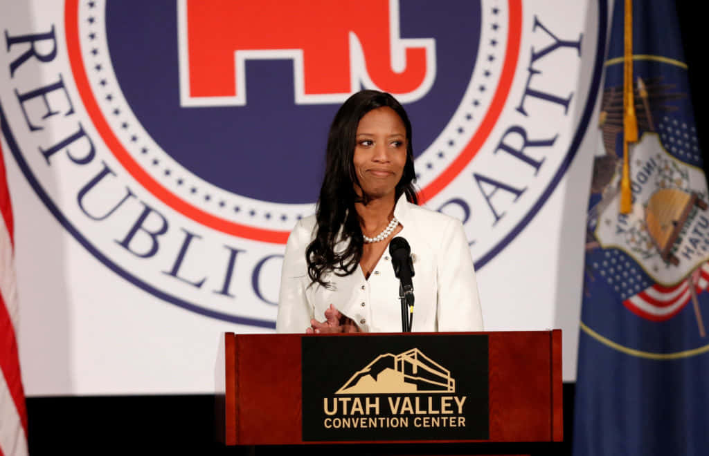 政治 桌布 壁紙 — 一位身穿白色西裝外套的女性站在印有「Utah Valley Convention Center」標誌的講台上，在共和黨背景和旗幟前發言。