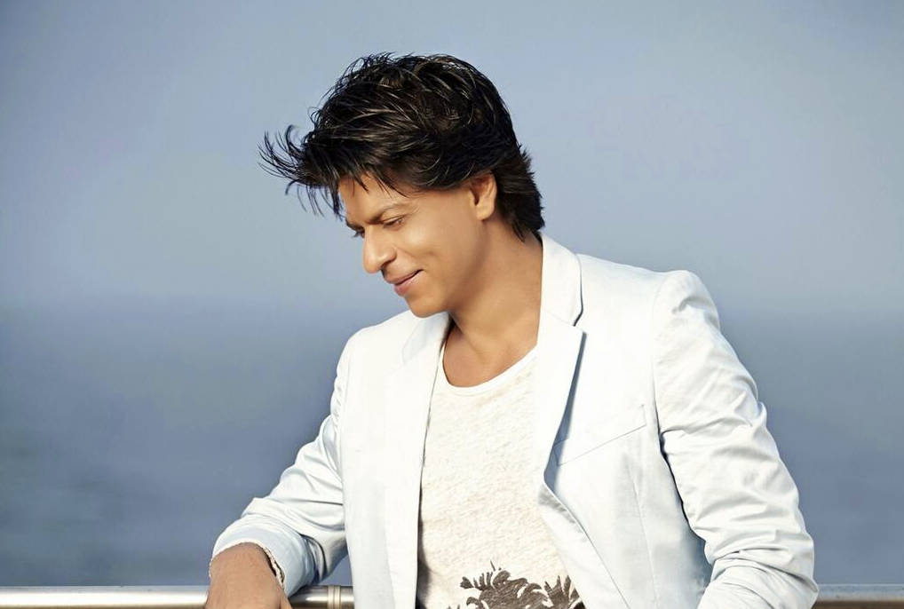Shahrukh 桌布 壁紙 — 一位身穿白色西裝外套的男子，在柔和的藍色背景下，靠著欄杆溫柔地微笑。