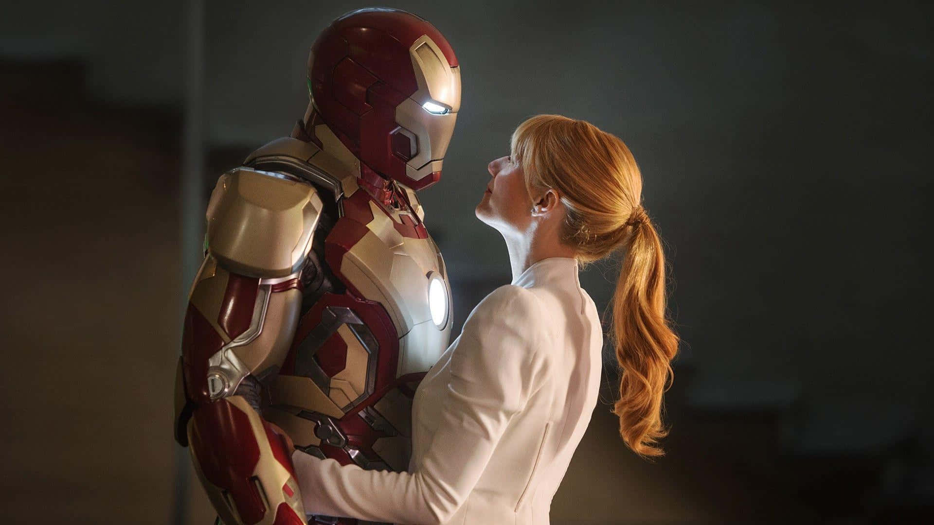 Pepper Potts 桌布 壁紙 — 一位身穿白色西裝的女性在昏暗的房間裡擁抱鋼鐵人，柔和的光線突顯了他們親密的時刻。