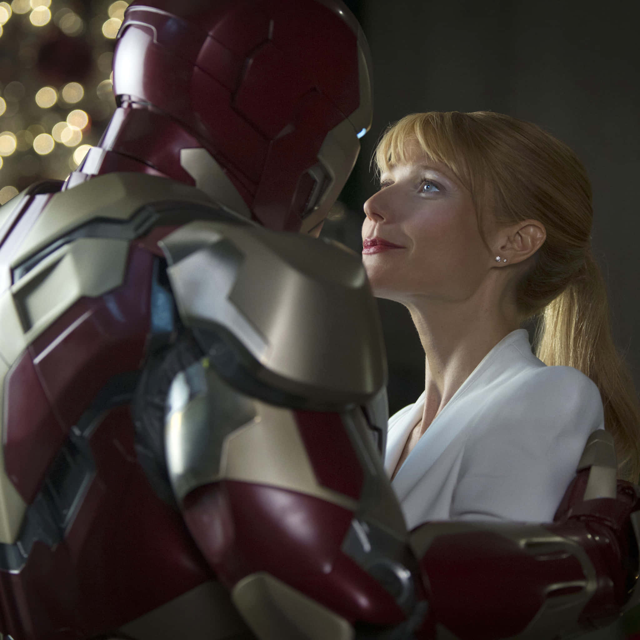 Pepper Potts 桌布 壁紙 — 一位身穿白色西裝的女性在溫暖的室內場景中，抬頭仰望鋼鐵人的紅色和金色裝甲，背景有散景燈光。