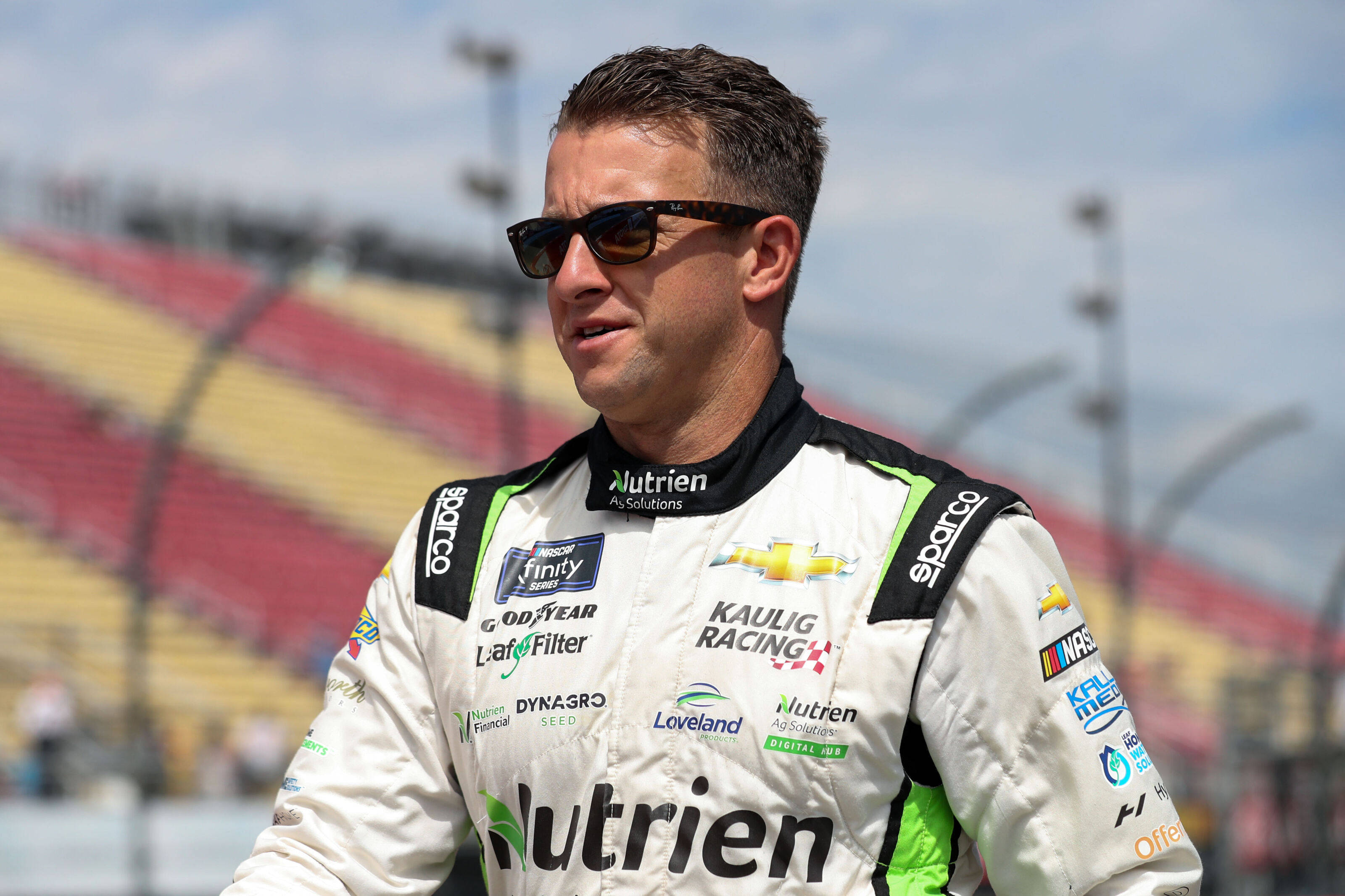 Man 桌布 壁紙 — 一位身穿白色賽車服、帶有贊助商標誌的NASCAR車手自信地站在賽道上，身後是空無一人的紅色和黃色看台。