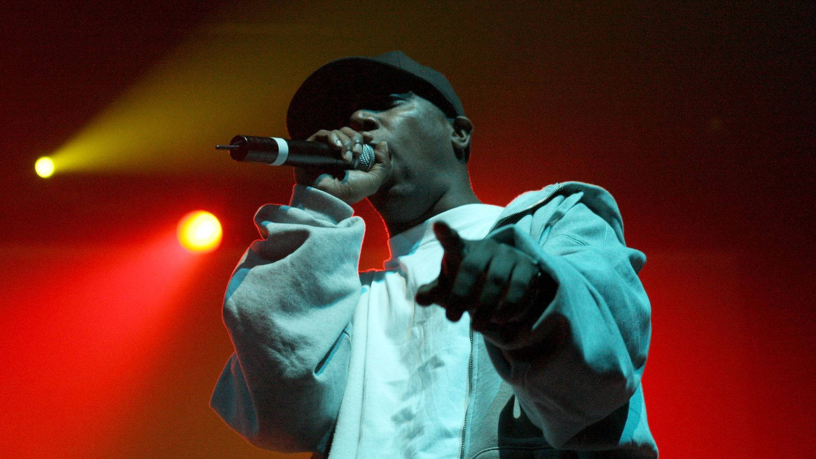 90s Rapper 桌布 壁紙 — 一位身穿白色連帽衫和黑色帽子的饒舌歌手在充滿活力的紅色和黃色舞台燈光下熱情表演。