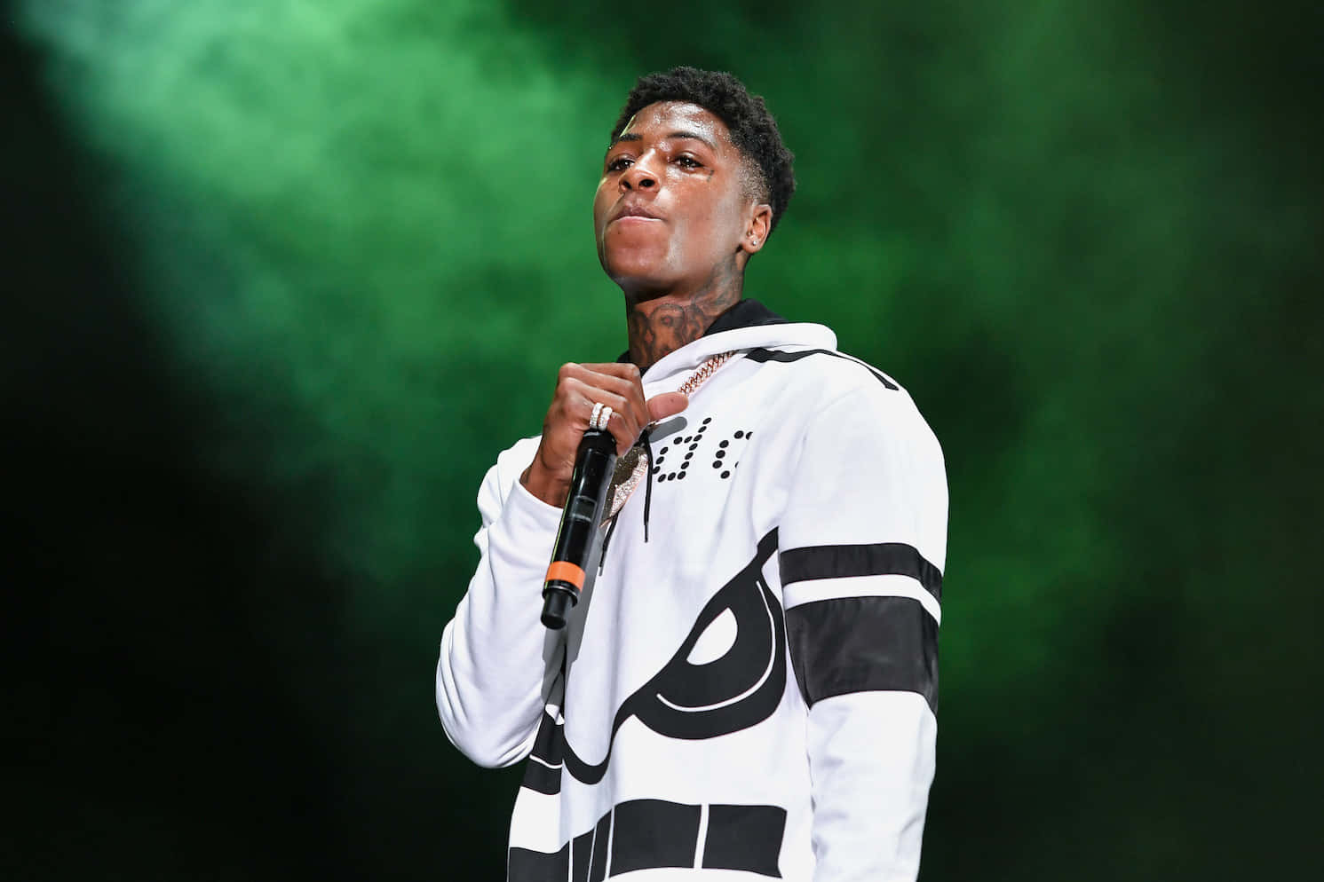 Youngboy 桌布 壁紙 — 一位身穿白色連帽衫的年輕男說唱歌手在綠色煙霧燈光下拿著麥克風在舞台上表演。