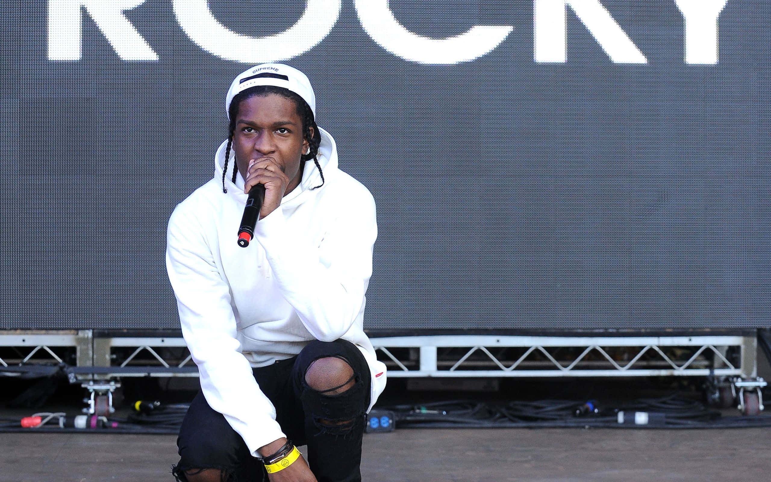 Asap Rocky 桌布 壁紙 — 一位身穿白色連帽衫的饒舌歌手跪在舞台上，在大型「ROCKY」螢幕下拿著麥克風。
