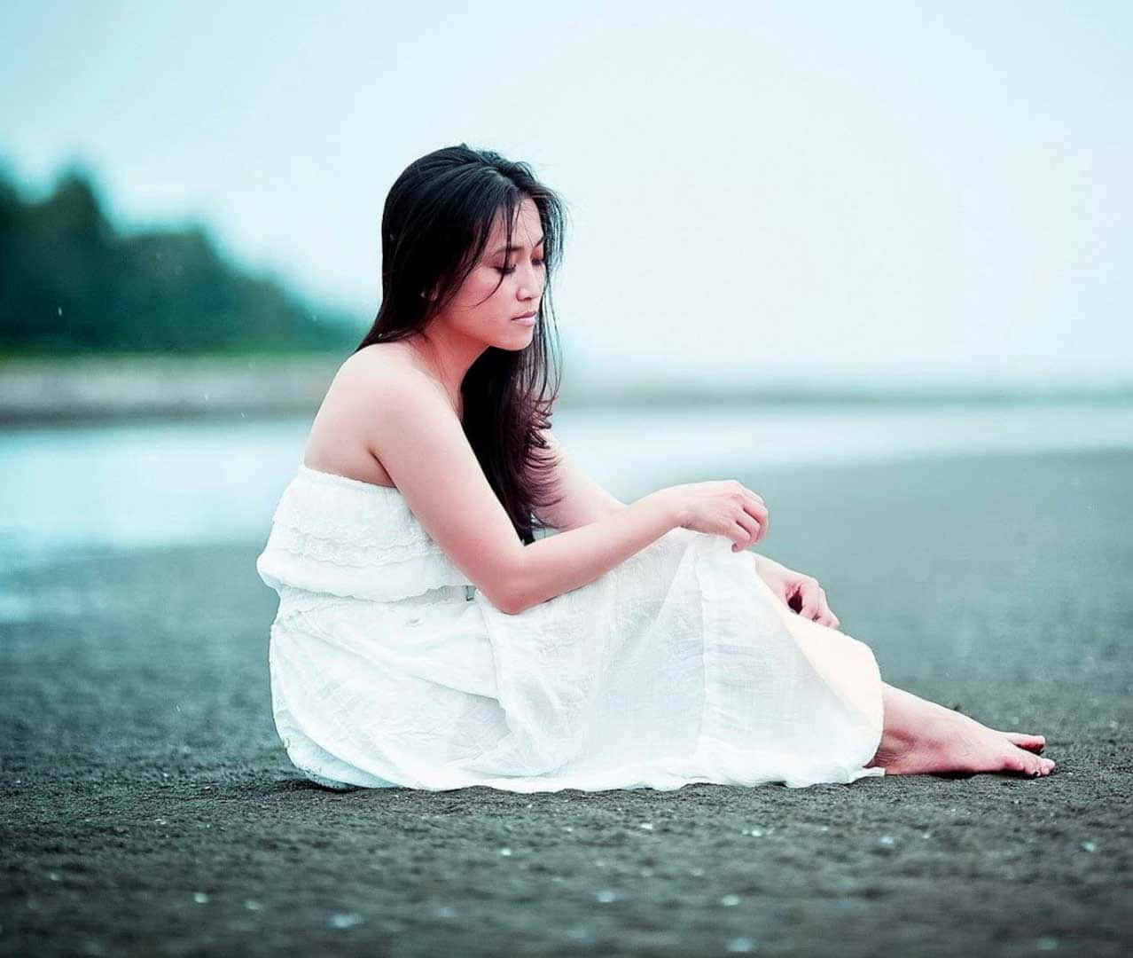 Depressing Profile 桌布 壁紙 — 一位身穿白色連衣裙的女子赤腳坐在黑暗的海灘上，低頭凝視，表情平靜，雨水輕輕落在她周圍。