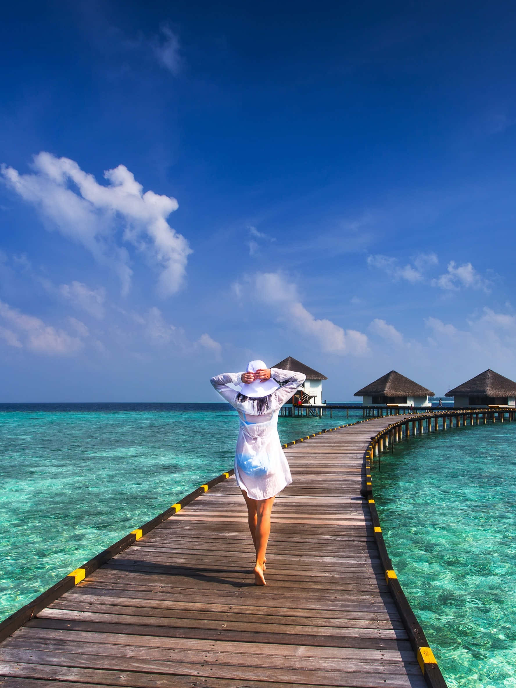 Travel Desktop 桌布 壁紙 — 一位身穿白色連衣裙的女子赤腳走在木製碼頭上，腳下是碧綠的海水，朝著水上平房走去，頭頂是明亮的藍天。
