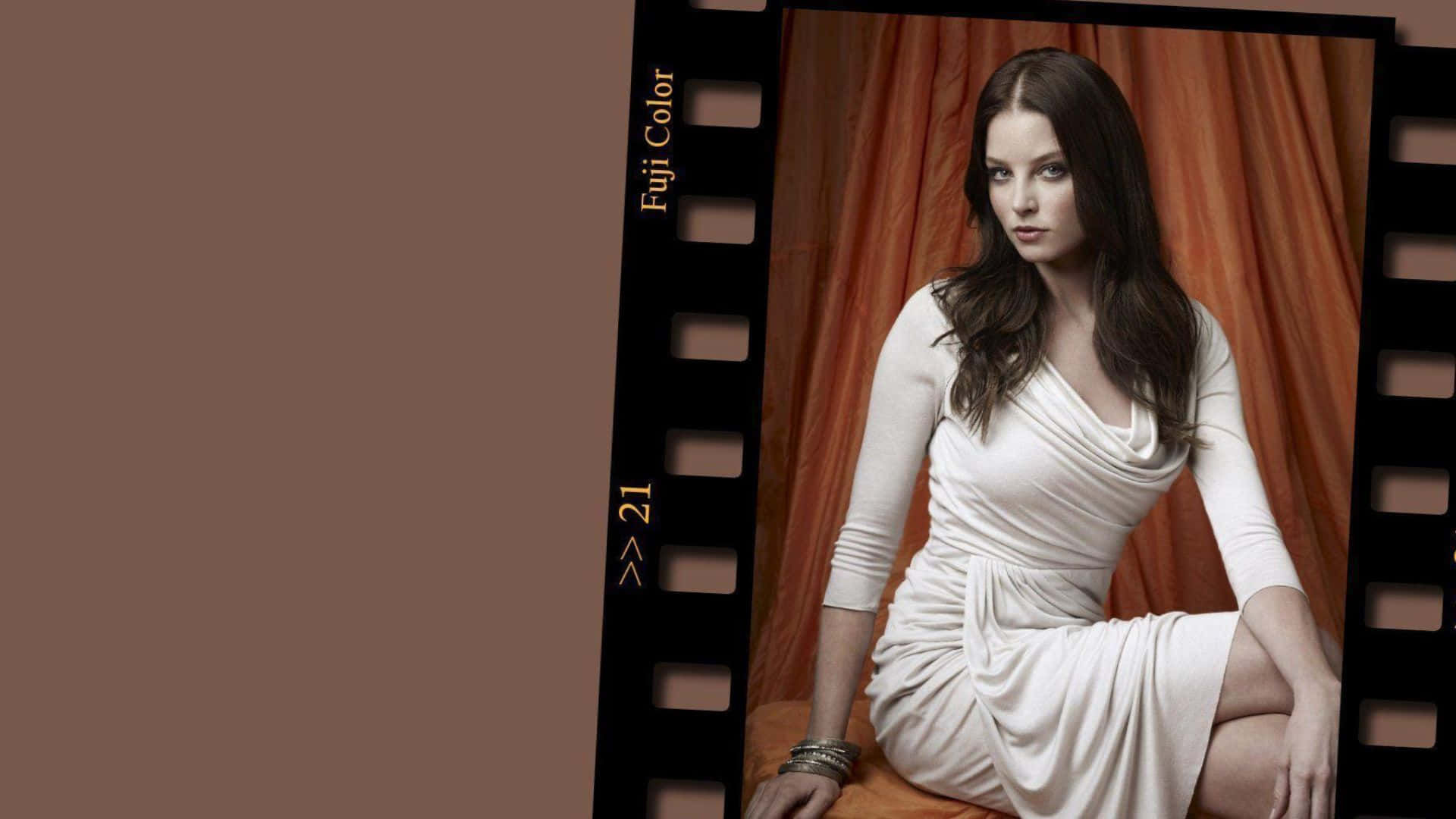 rachel nichols 桌布 壁紙 — 一位身穿白色連衣裙的女性優雅地坐在溫暖的橙色窗簾前，窗簾被黑色膠卷邊框框住。