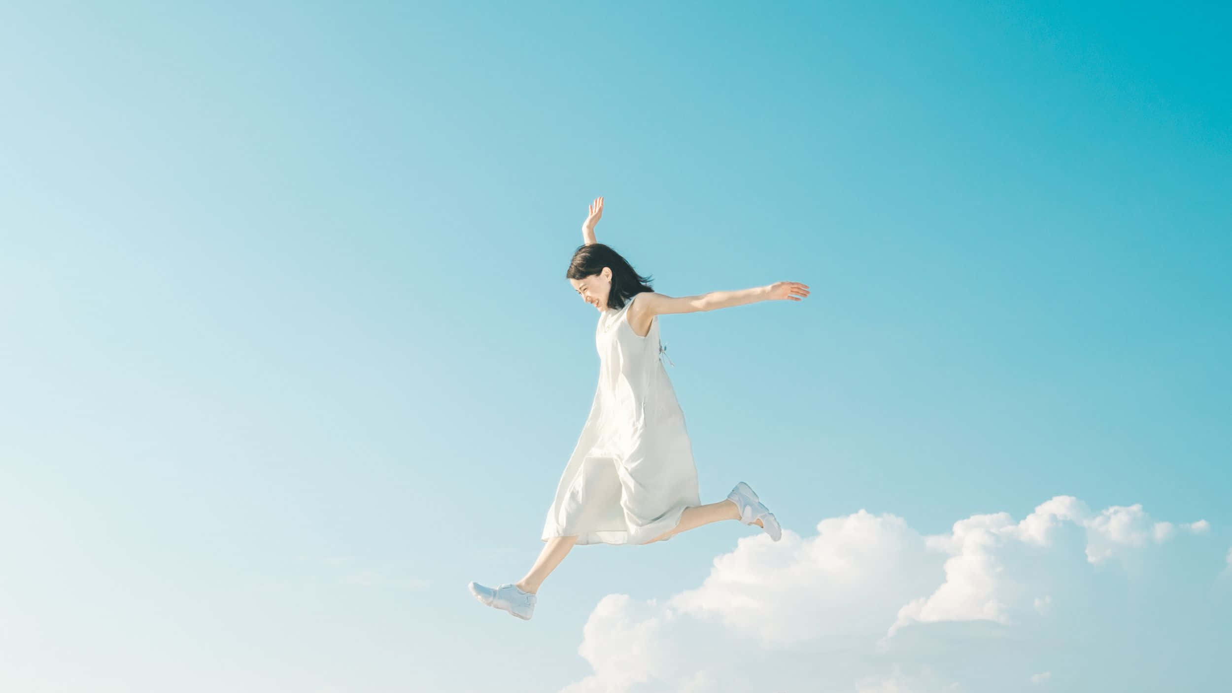 jump 桌布 壁紙 — 一位身穿白色連衣裙的女性在清澈的藍天和下方蓬鬆的雲朵中歡快地跳躍。
