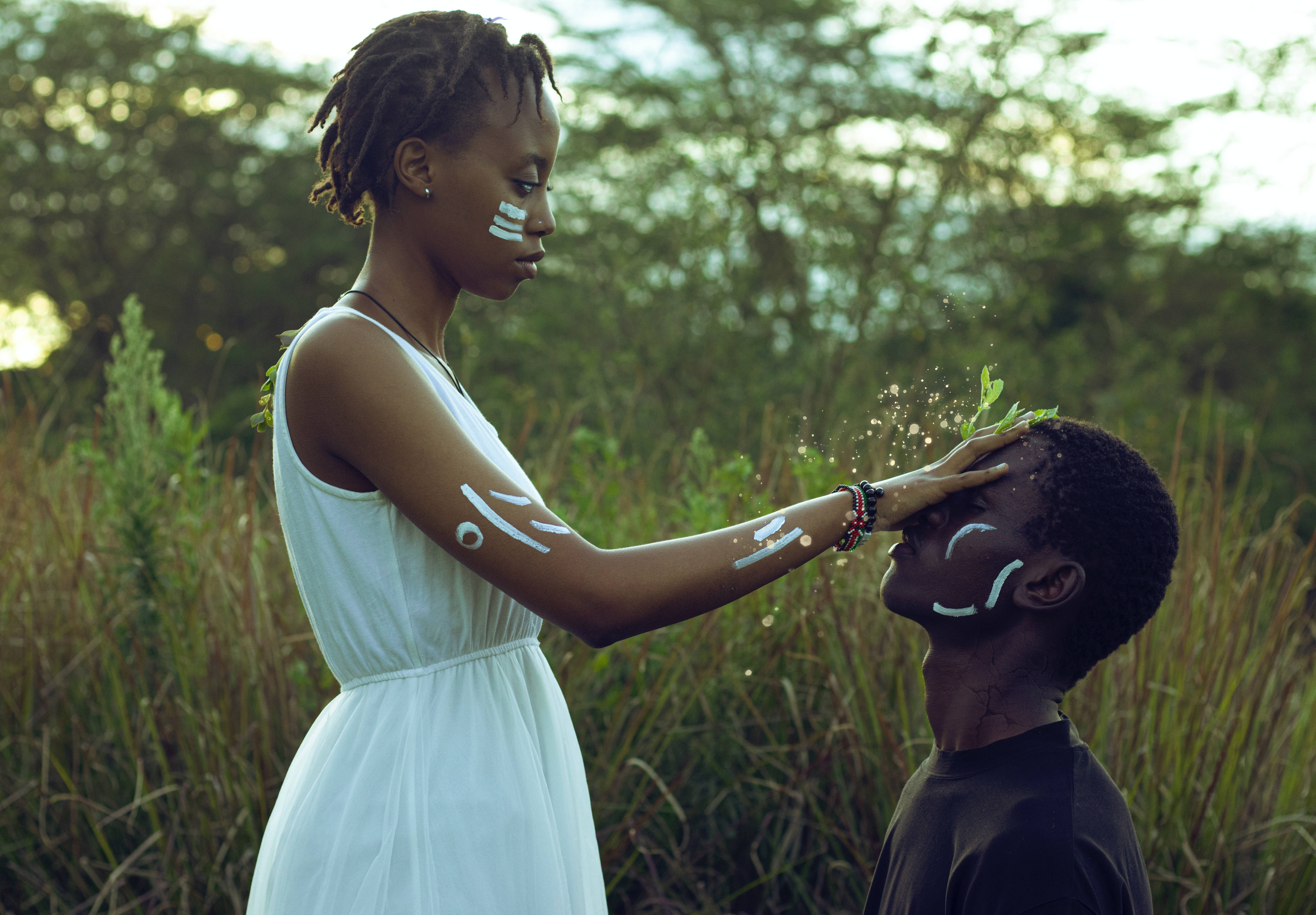 Black Couple 桌布 壁紙 — 一位身穿白色連衣裙的女性輕輕地將手放在一個孩子的頭上，綠葉和發光的粒子在陽光照耀的田野中漂浮在他們周圍。