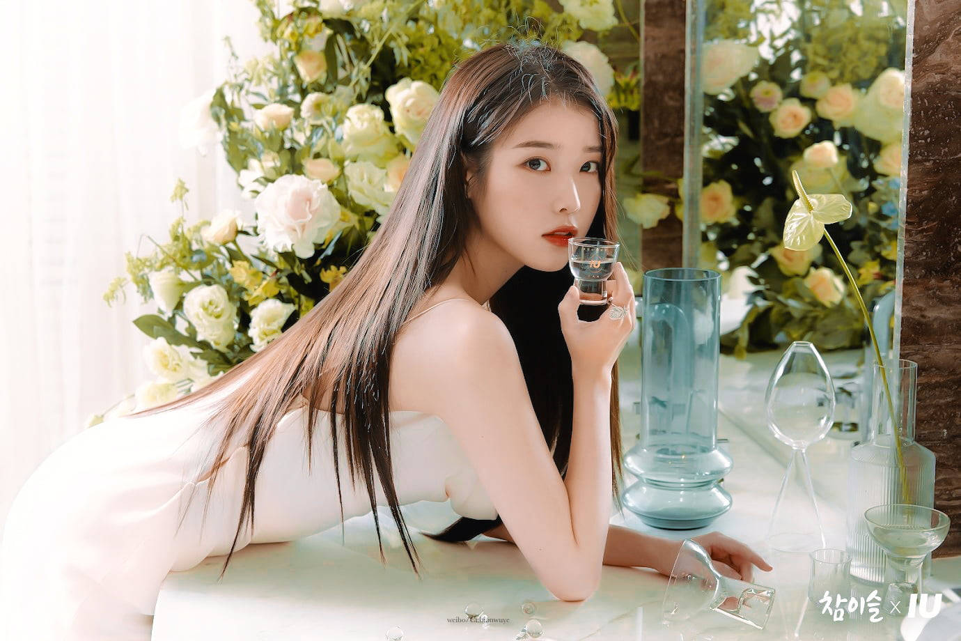 Magazine 桌布 壁紙 — 一位身穿白色連衣裙的女性靠在桌子上，手持酒杯，周圍環繞著茂盛的花卉佈置和優雅的玻璃器皿。