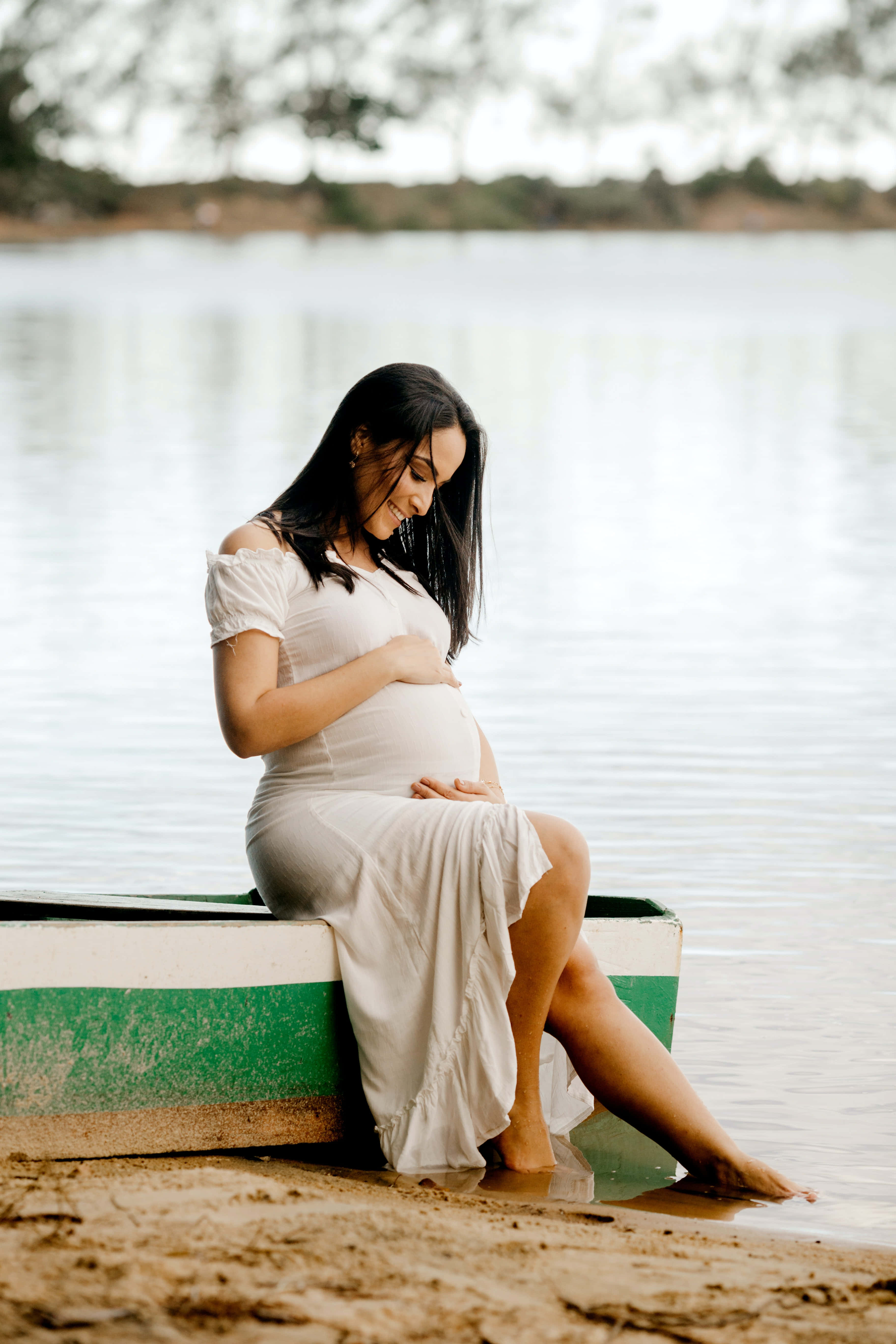 Pregnant 桌布 壁紙 — 一位身穿白色連衣裙的孕婦，靜靜地坐在平靜湖面上的一艘綠色和白色的小船上，帶著平靜的微笑撫摸著她的肚子。