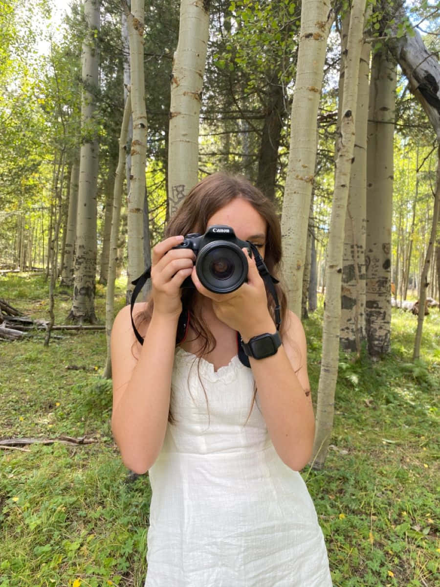 Holding Camera 桌布 壁紙 — 一位身穿白色連衣裙的年輕女子站在陽光普照的森林中高大的白楊樹之間，將相機舉到臉前。