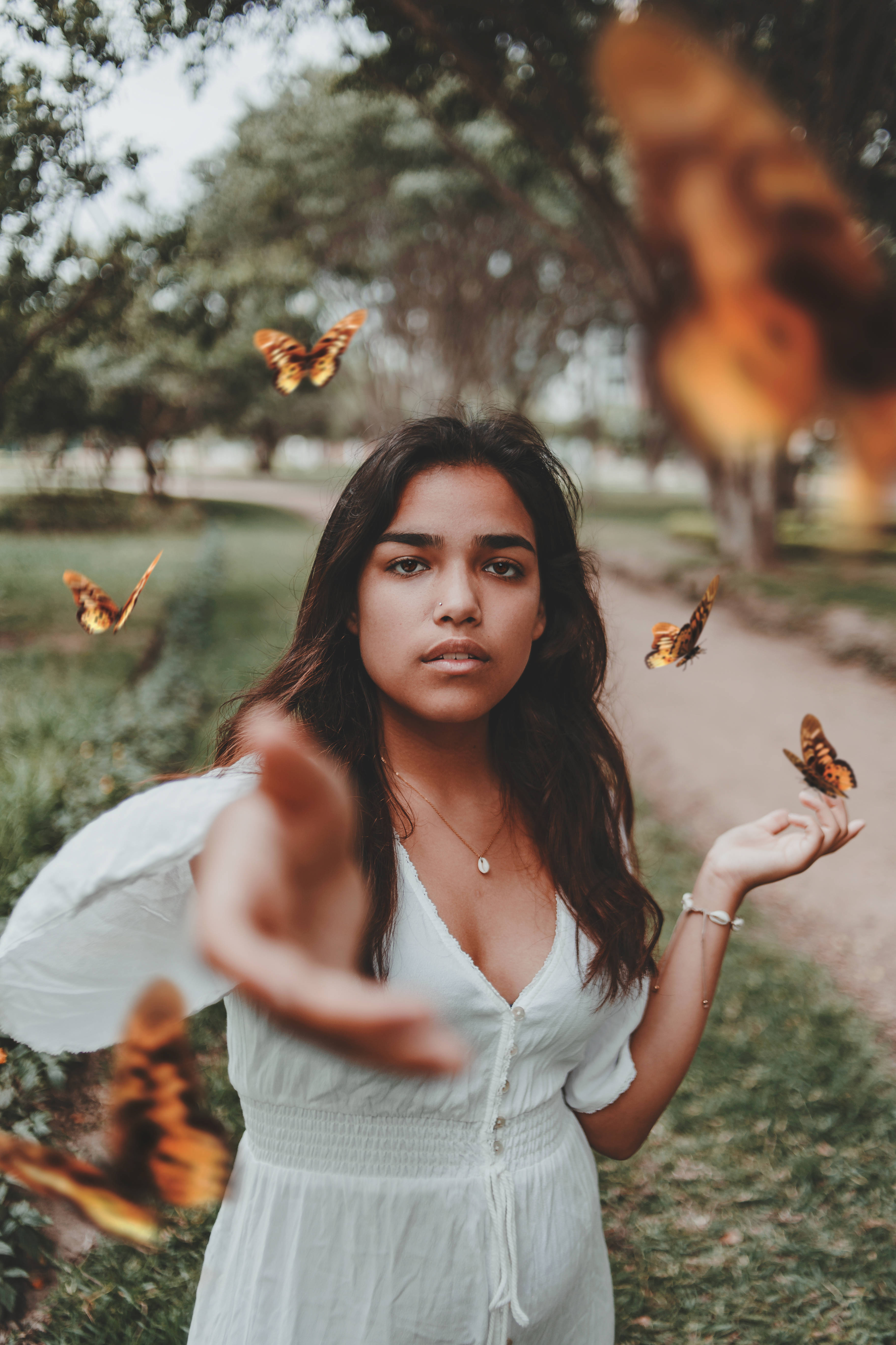 Cute Butterfly 桌布 壁紙 — 一位身穿白色連衣裙的年輕女子站在陽光明媚的公園裡，橙色蝴蝶在她手和臉周圍翩翩起舞。