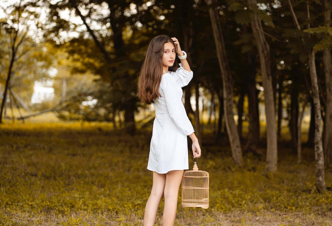 Teen Girl 桌布 壁紙 — 一位身穿白色連衣裙的年輕女子站在陽光明媚的森林中，手持柳條鳥籠，周圍是金色的樹葉。