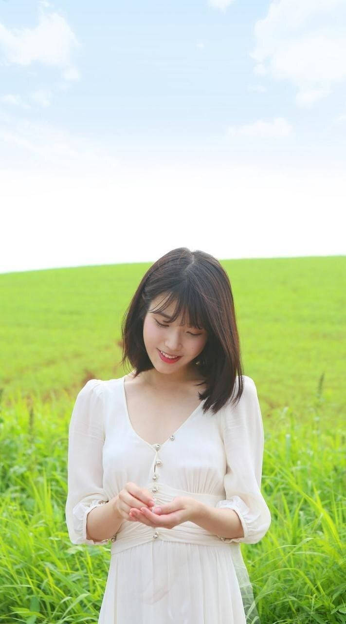 iu phone 桌布 壁紙 — 一位身穿白色連衣裙的年輕女性在晴朗藍天下，於陽光明媚的綠色田野中輕握著草，溫柔微笑。