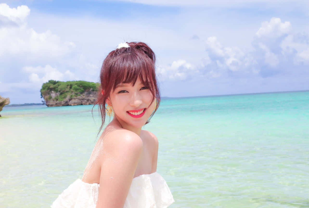twice 桌布 壁紙 — 一位身穿白色連衣裙的快樂女士在陽光明媚的海灘上對著鏡頭微笑，背景是碧綠的海水和一個岩石島嶼。