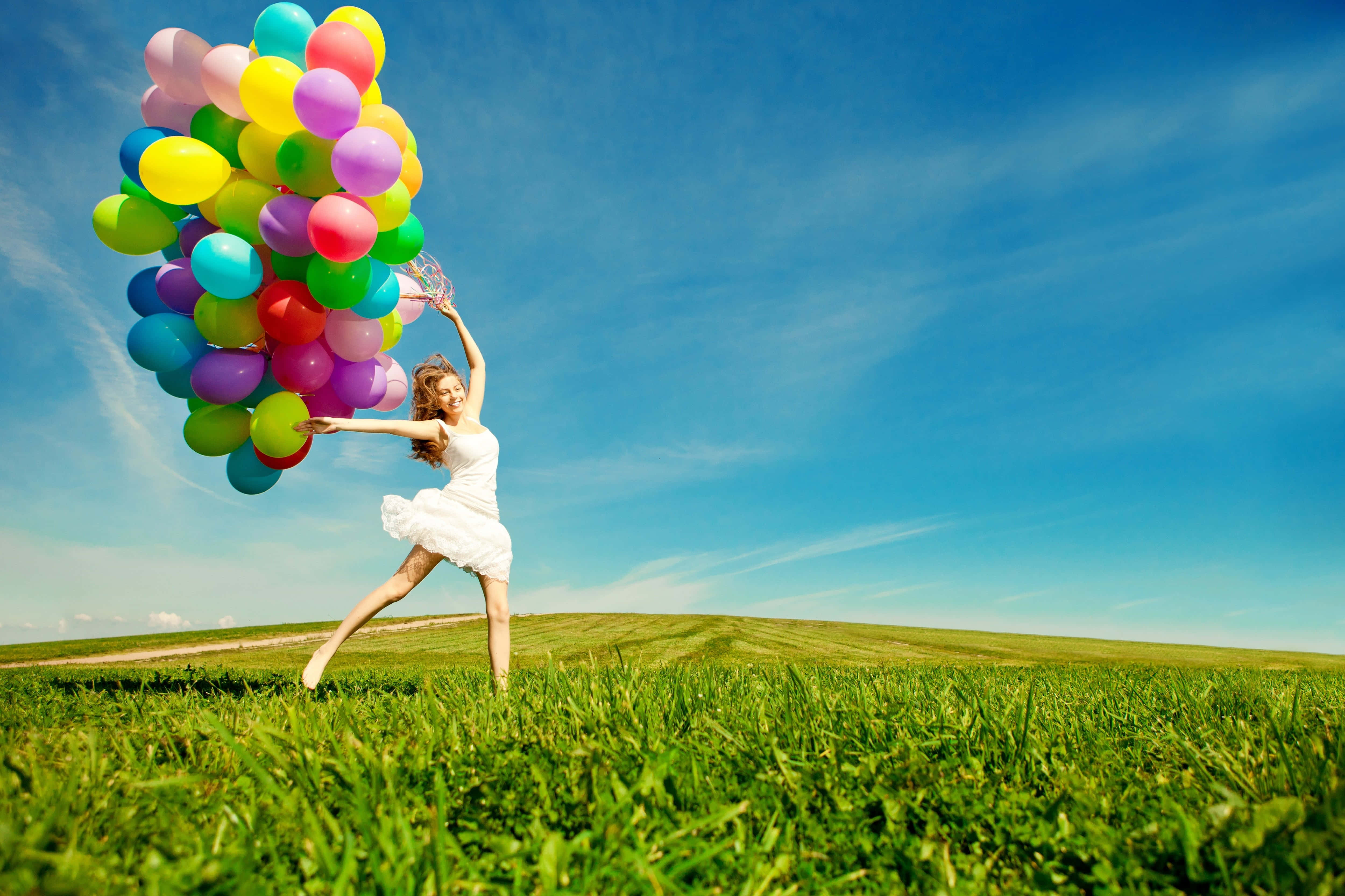 Balloon 桌布 壁紙 — 一位身穿白色連衣裙的快樂女性，在充滿活力的綠色田野中跳舞，手持一大束彩虹色氣球，背景是晴朗的藍天。