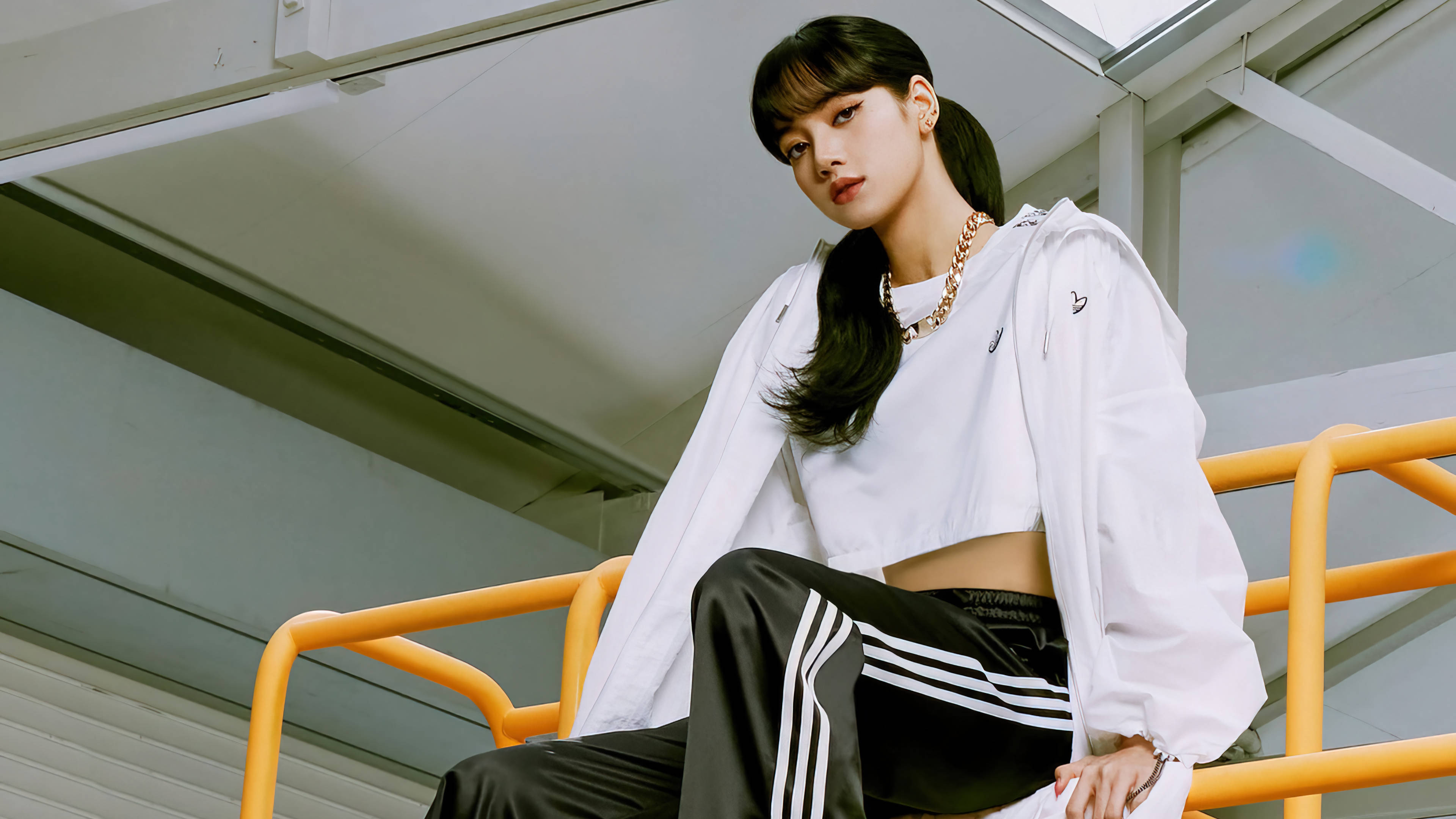lisa 桌布 壁紙 — 一位身穿白色運動服的年輕女性自信地坐在工業風格燈光下的亮橙色欄杆上。