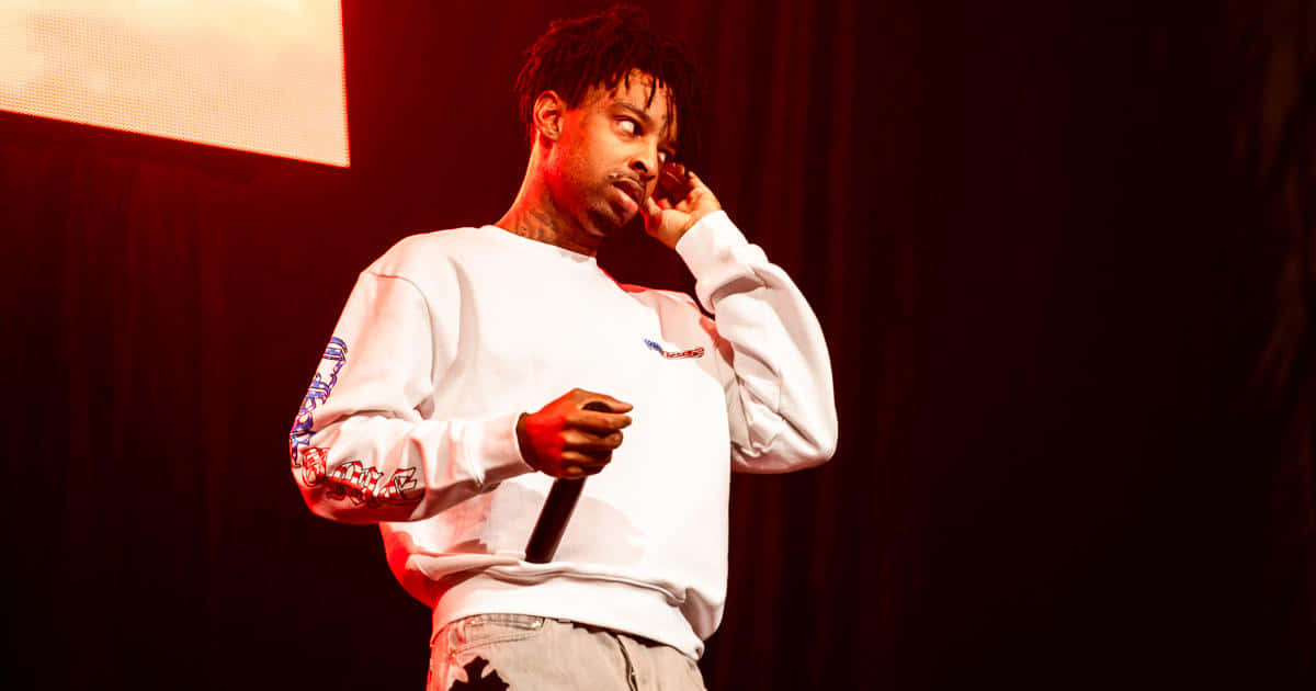 21 Savage 桌布 壁紙 — 一位身穿白色運動衫的男性饒舌歌手在溫暖的紅色舞台燈光下，以深色窗簾背景為襯，手持麥克風。