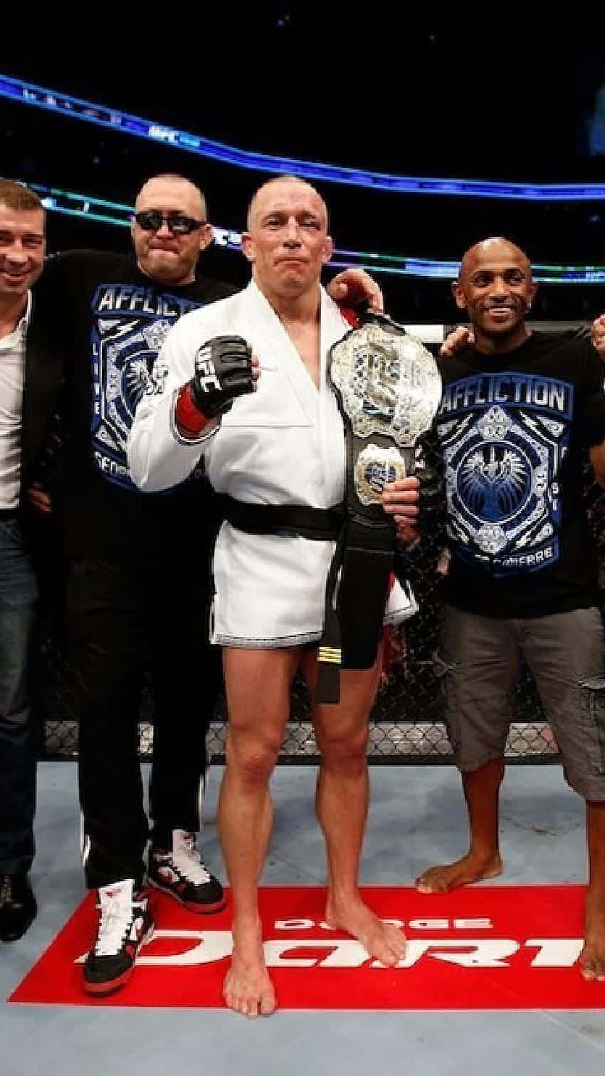 martial arts 桌布 壁紙 — 一位身穿白色道服的勝利混合武術選手，赤腳站在紅色墊子上，與隊友在燈光明亮的八角籠中高舉冠軍腰帶。