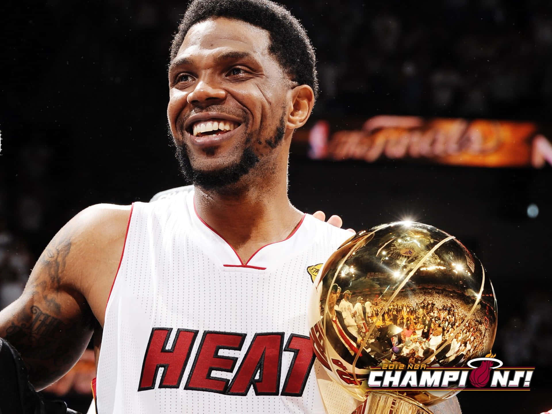 Udonis Haslem 桌布 壁紙 — 一位身穿白色邁阿密熱火球衣的勝利籃球運動員，面帶笑容，手持閃閃發光的2012年NBA總冠軍獎盃。