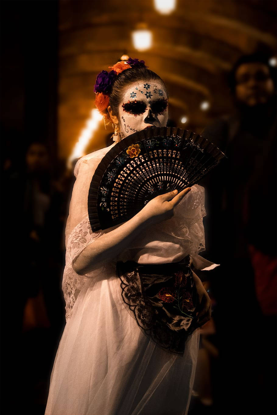 Santa Muerte 桌布 壁紙 — 一位身穿白色長袍、化著糖骷髏妝容的女性，在溫暖的環境燈光下手持一把黑色扇子。