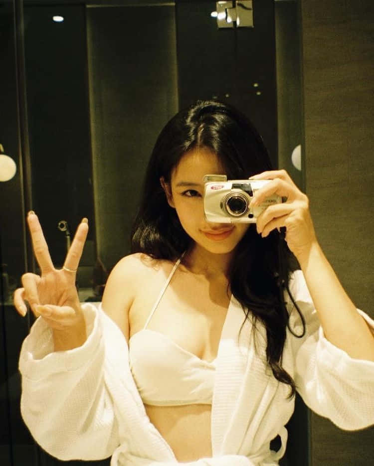 Holding Camera 桌布 壁紙 — 一位身穿白色長袍和辮子的女士微笑著用銀色相機和和平手勢在鏡子前自拍。
