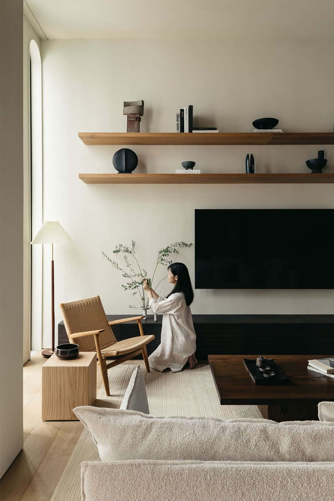 Minimalist Design 桌布 壁紙 — 一位身穿白色長袍的女性，在一個充滿柔和自然光線的極簡客廳中，將精緻的樹枝插在現代木椅旁的花瓶裡。