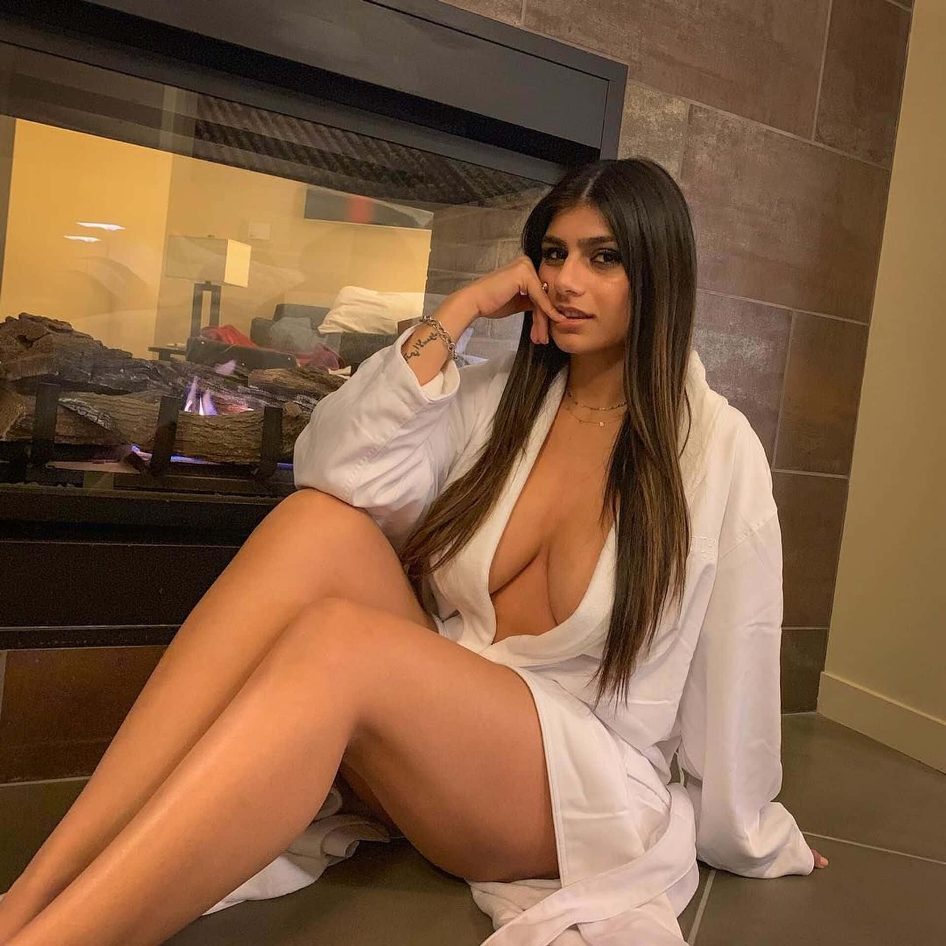 Mia Khalifa 桌布 壁紙 — 一位身穿白色長袍的女性優雅地坐在現代磁磚房間的發光壁爐前。
